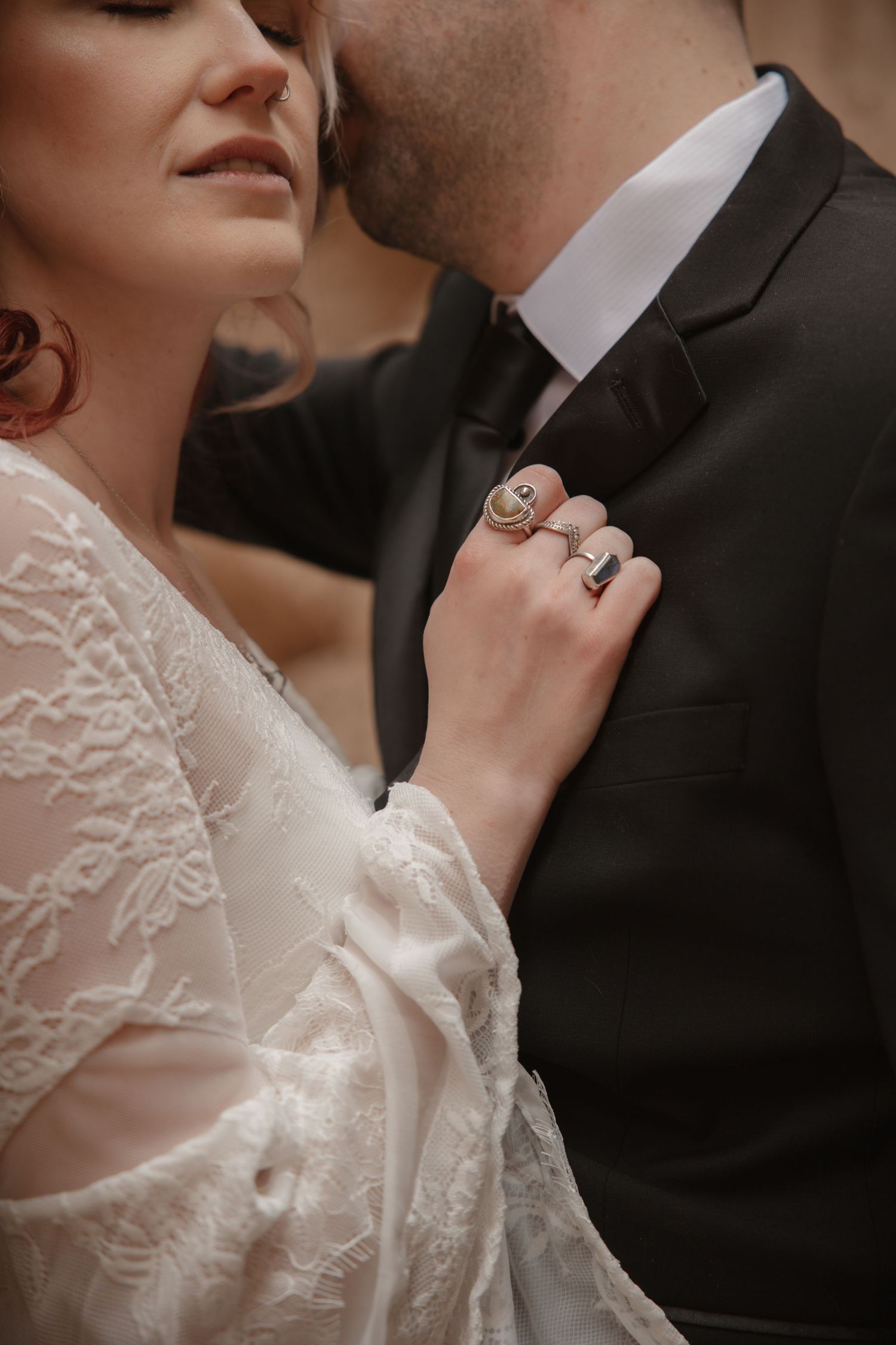 Close-up of brides hand with statement rings resting on grooms black tuxedo lapel during wedding.
