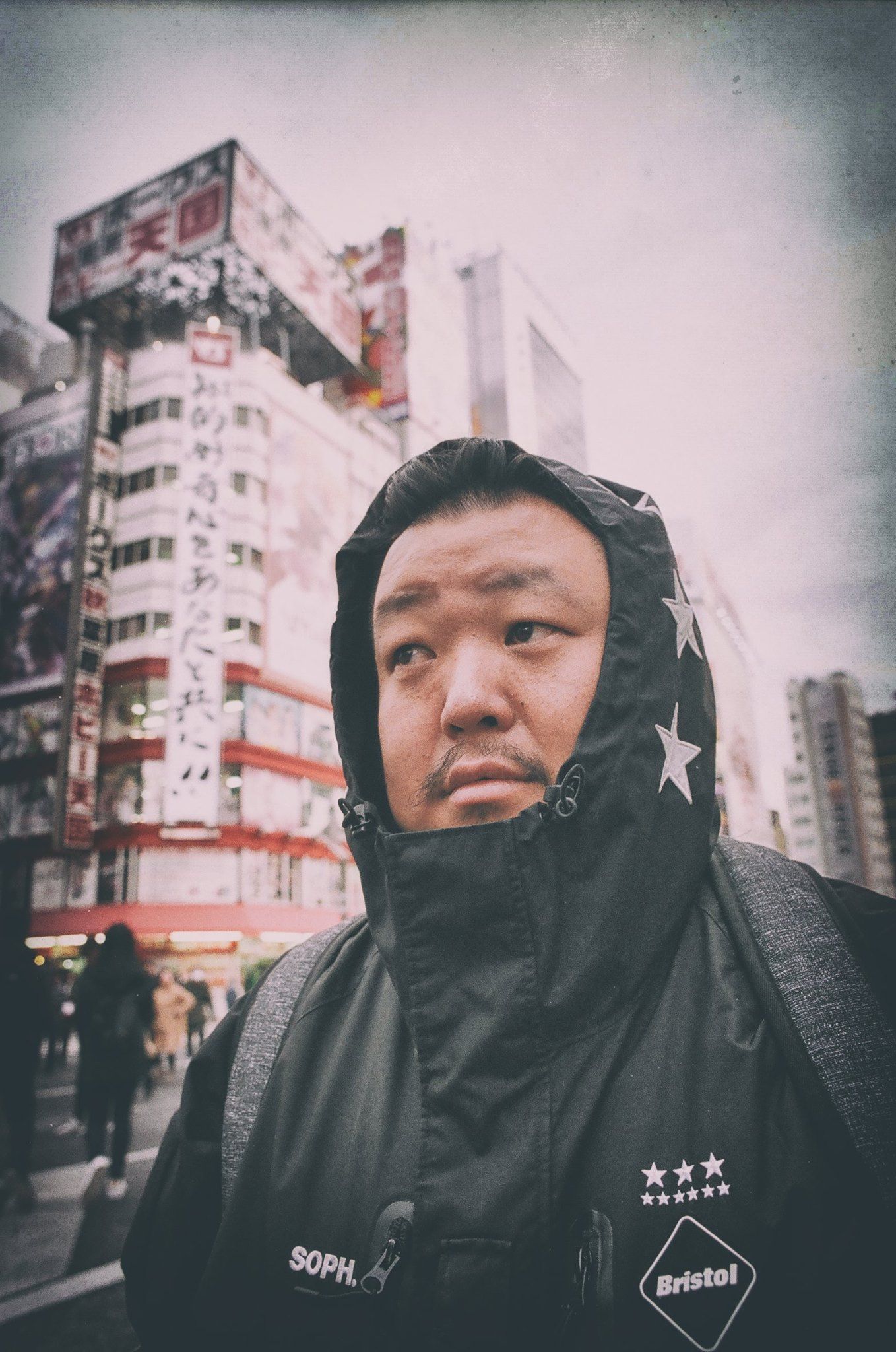 A young Asian man in a black hooded FC Bristol jacket stands on a busy urban street in Japan.