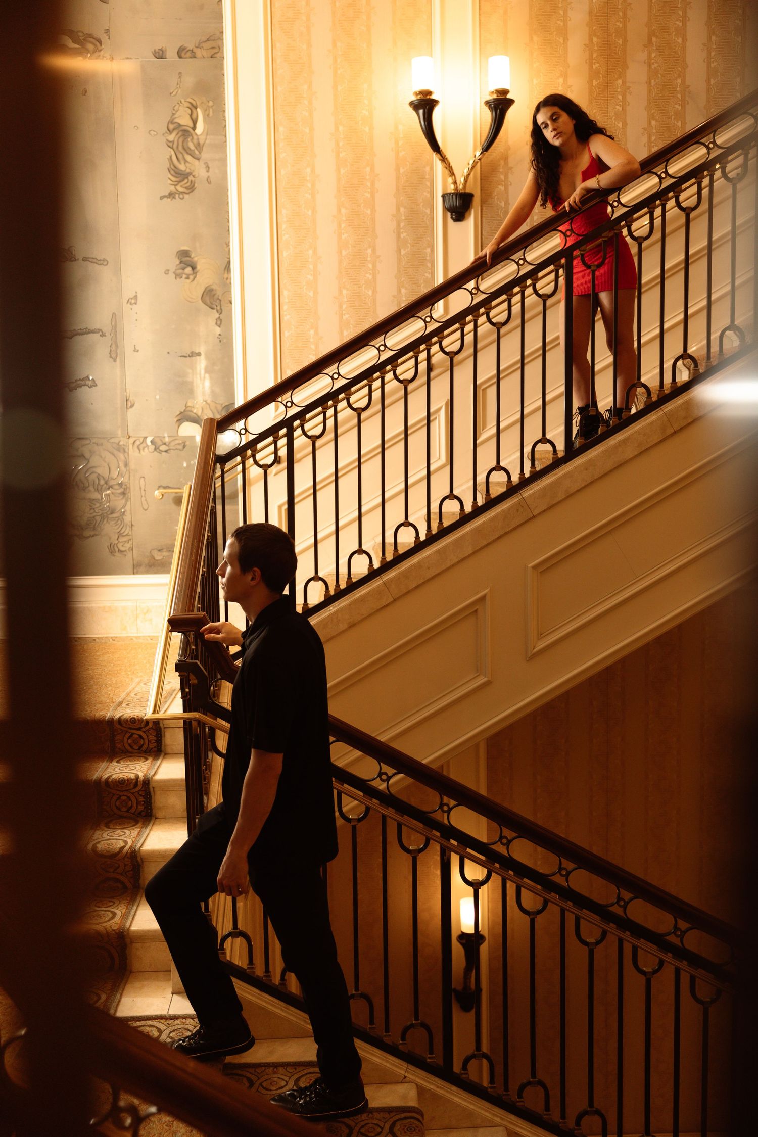 Couple on elegant staircase, woman in red dress stands above man in black shirt in luxury hotel interior.