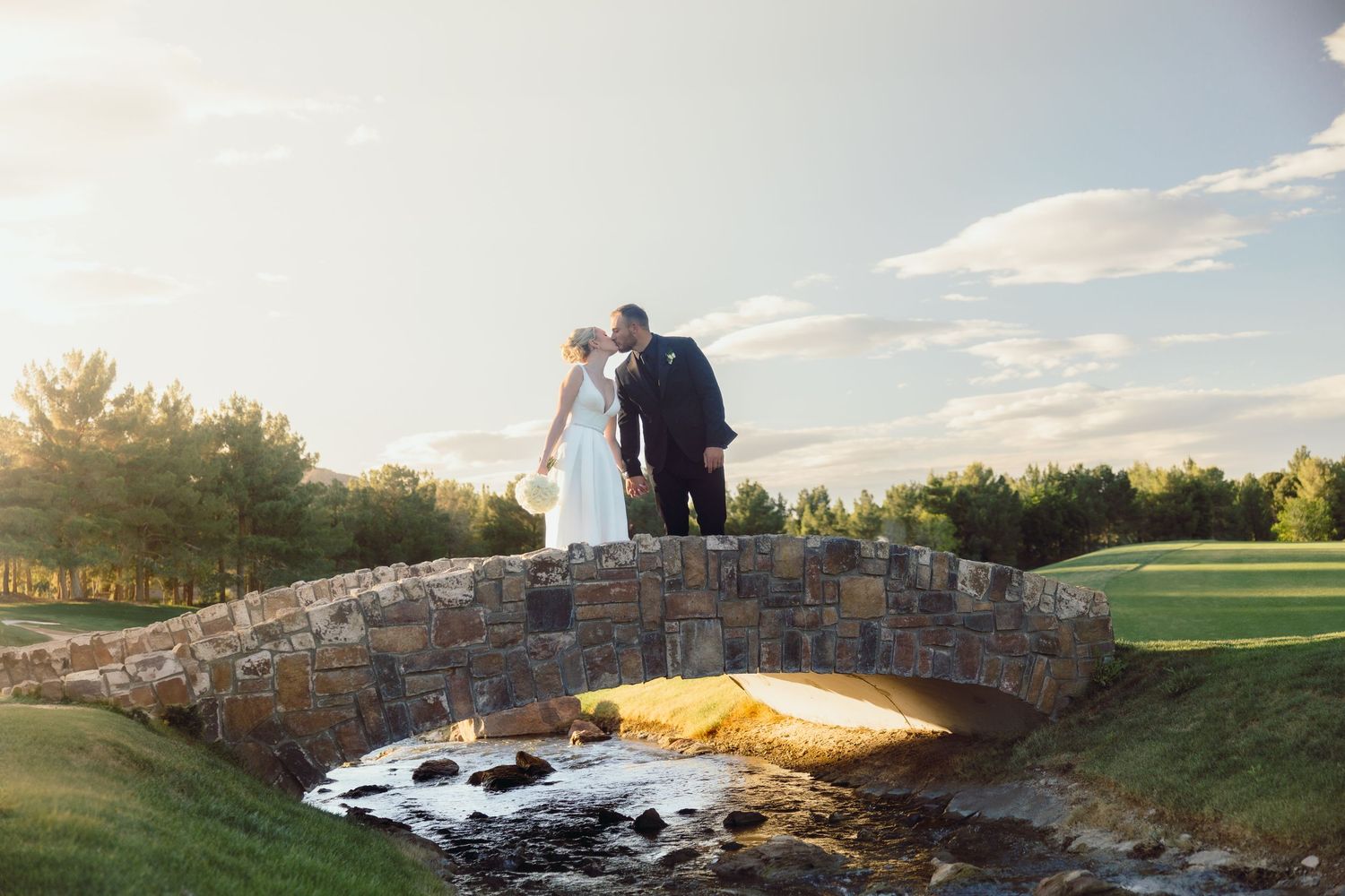 A bride and groom share a kiss on a rustic stone bridge at sunset on a scenic golf course.