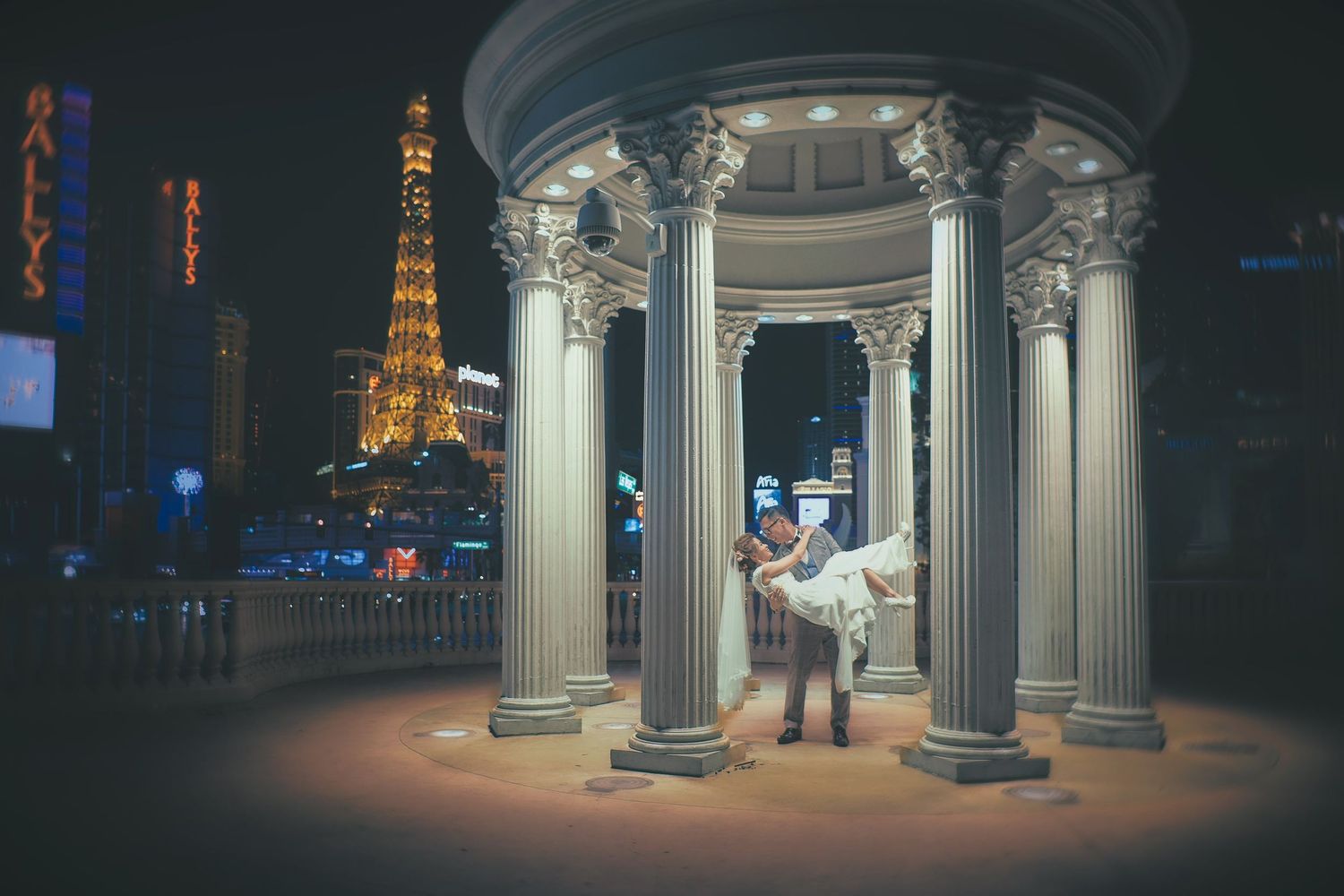 Couple embracing under Roman columns at night with Eiffel Tower replica on Las Vegas Strip glowing behind them.