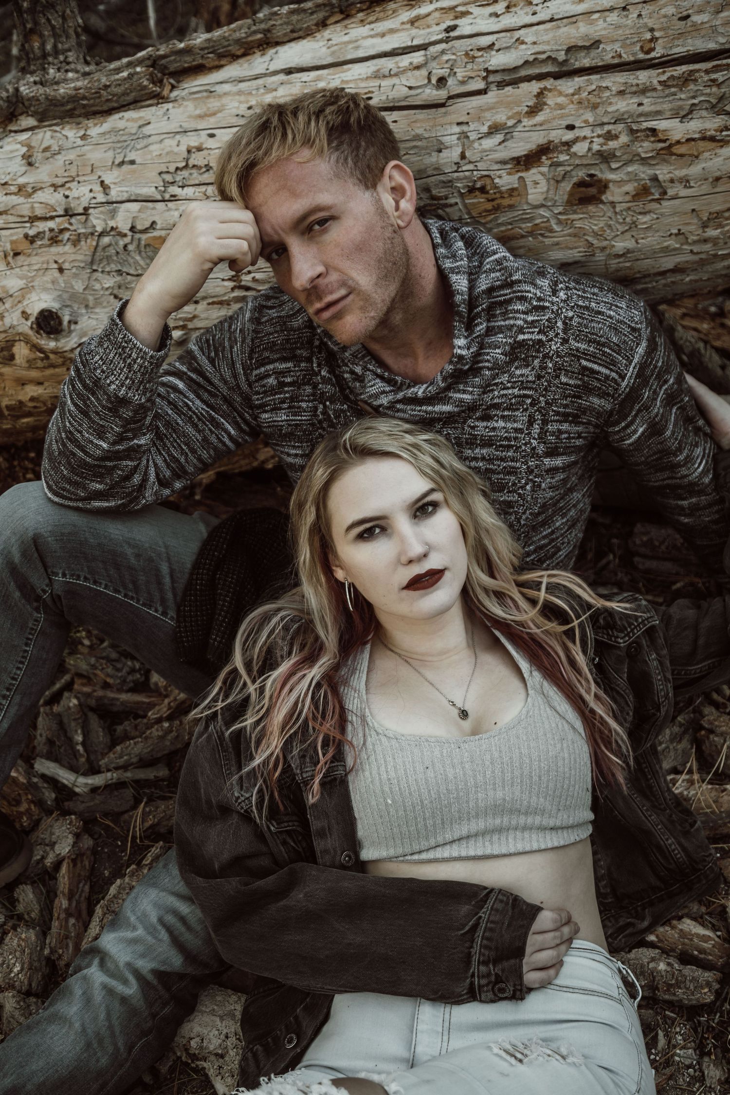 A couple poses against fallen logs outdoors, man in gray sweater behind woman in crop top and denim jacket.
