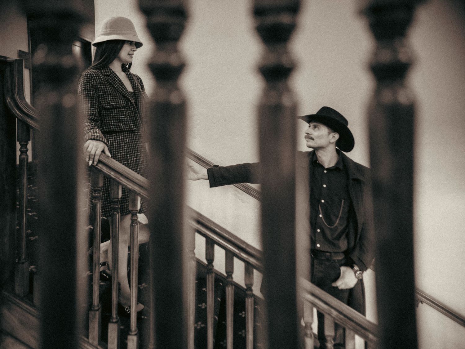 A man and woman in cowboy hats share a romantic moment on a vintage wooden staircase in sepia tone.