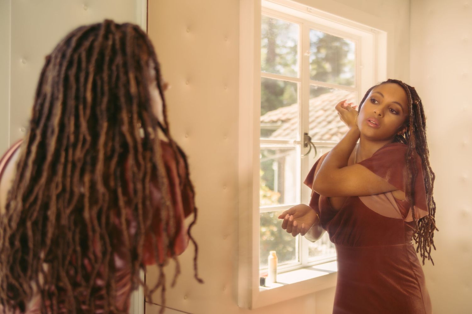 Woman with long red locs in rust dress adjusting hair in front of window mirror.