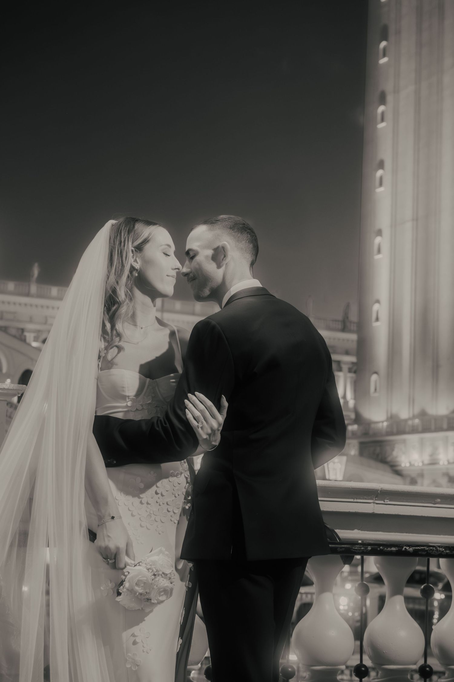 Black and white romantic portrait of a bride and groom nearly kissing on a balcony at night.