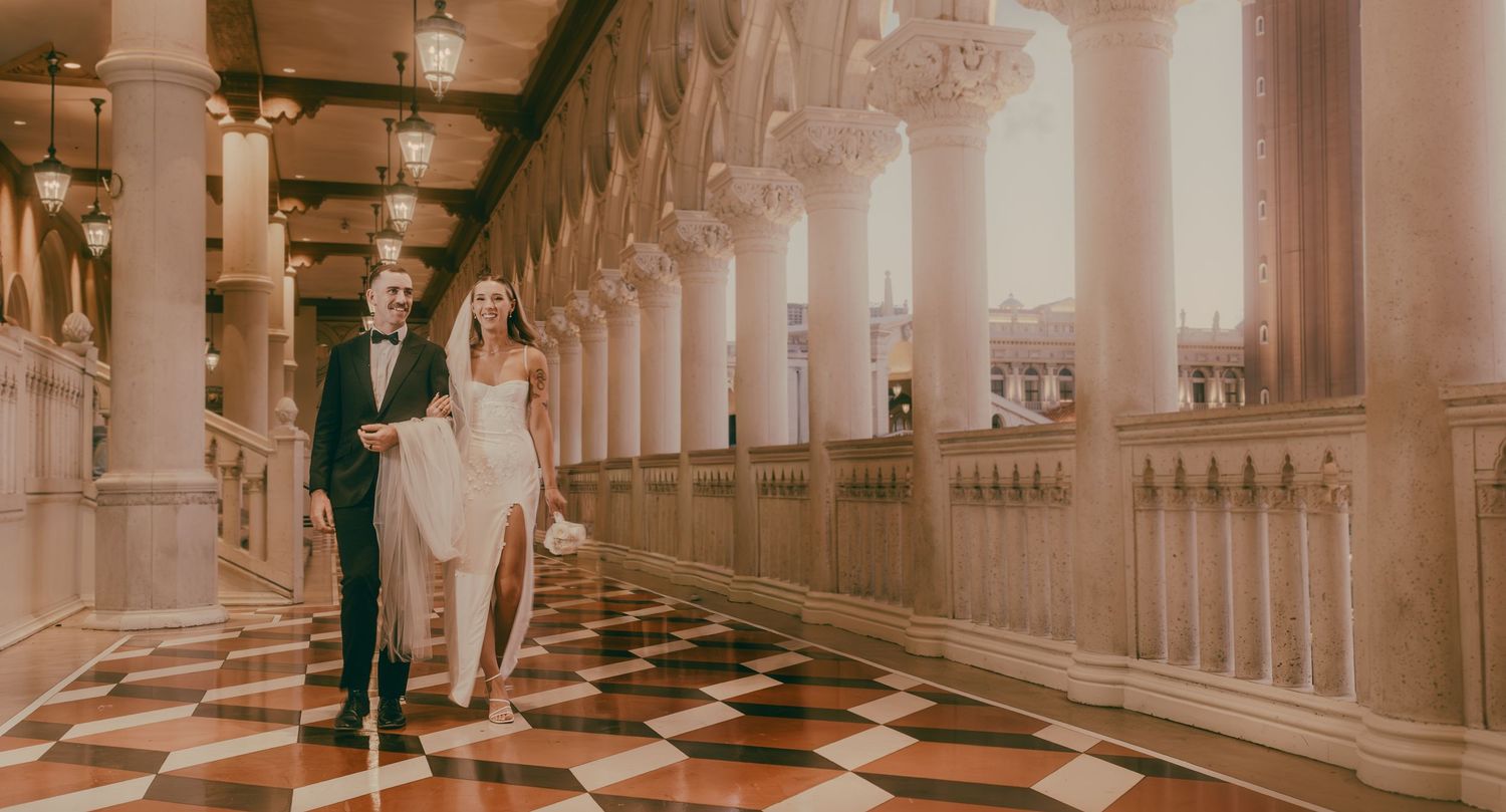 Bride and groom walk hand in hand through grand marble hallway with checkered floors.