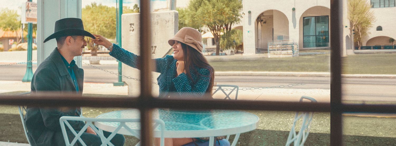 A couple sits at a teal outdoor table, woman adjusting man's black cowboy hat, in a sunny town setting.