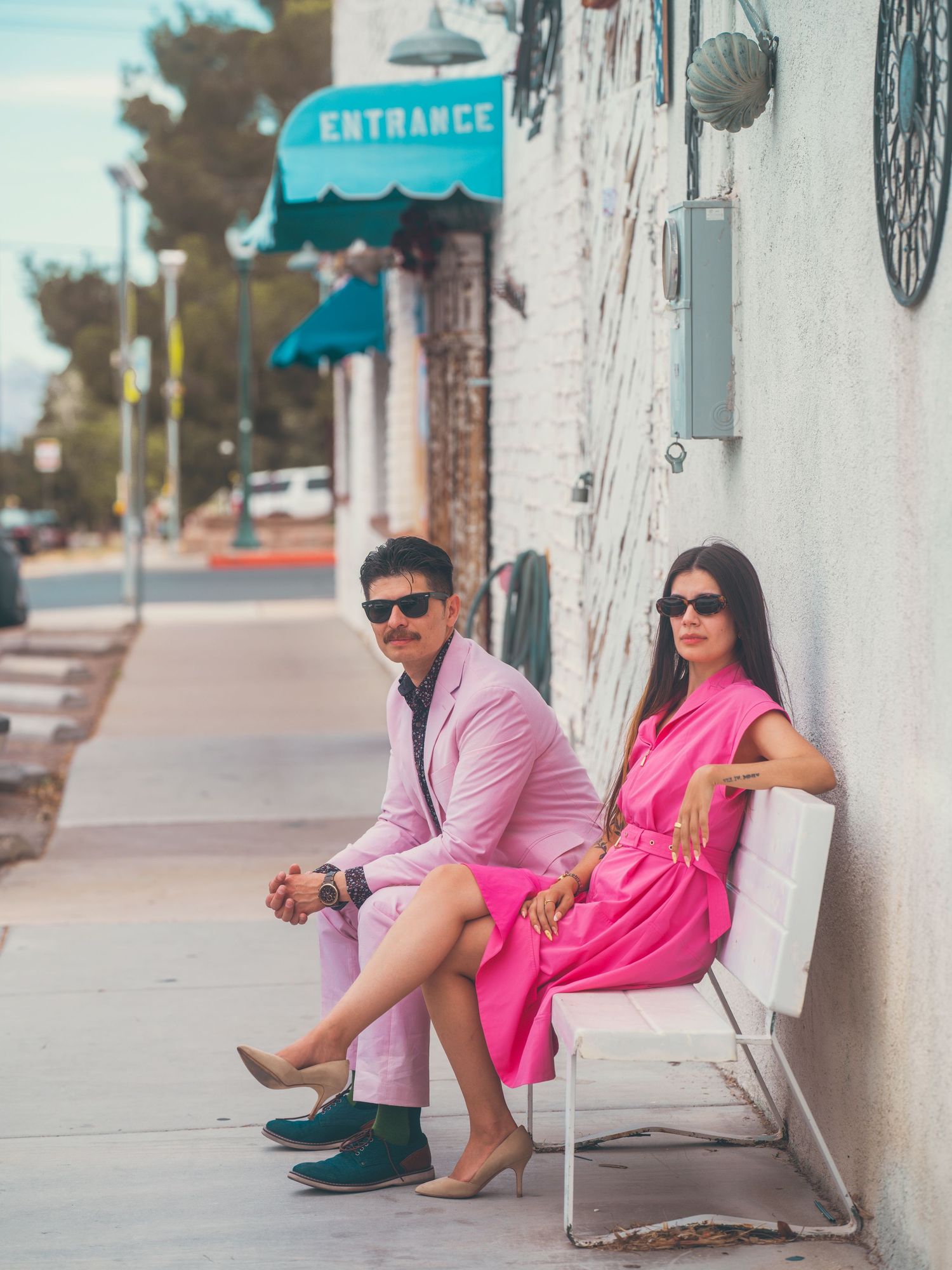 Stylish couple in pink outfits sitting outside a shop near an entrance sign on a sunny day.