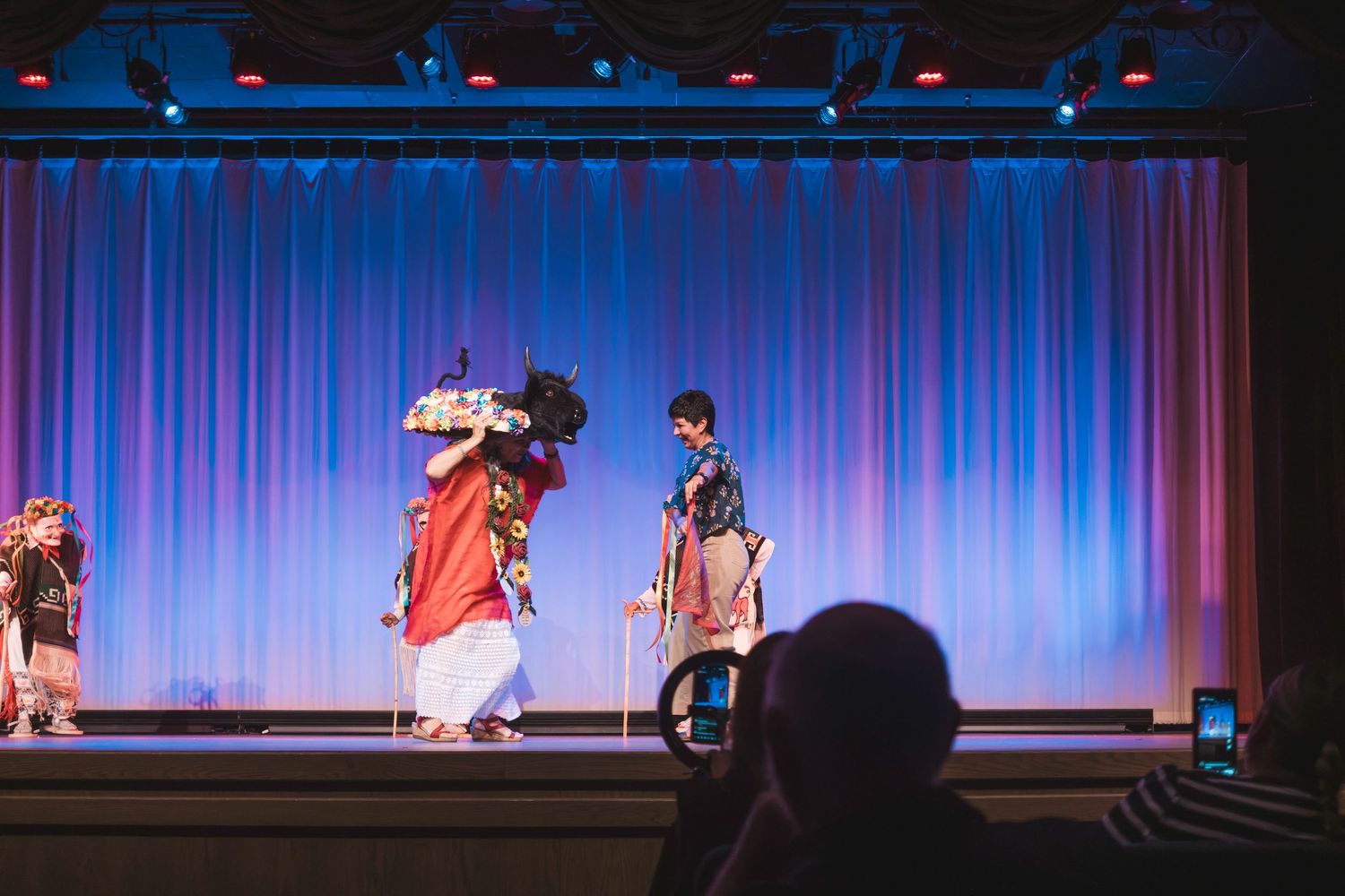 Performers in traditional costumes on a theater stage with blue curtains during a cultural dance performance.