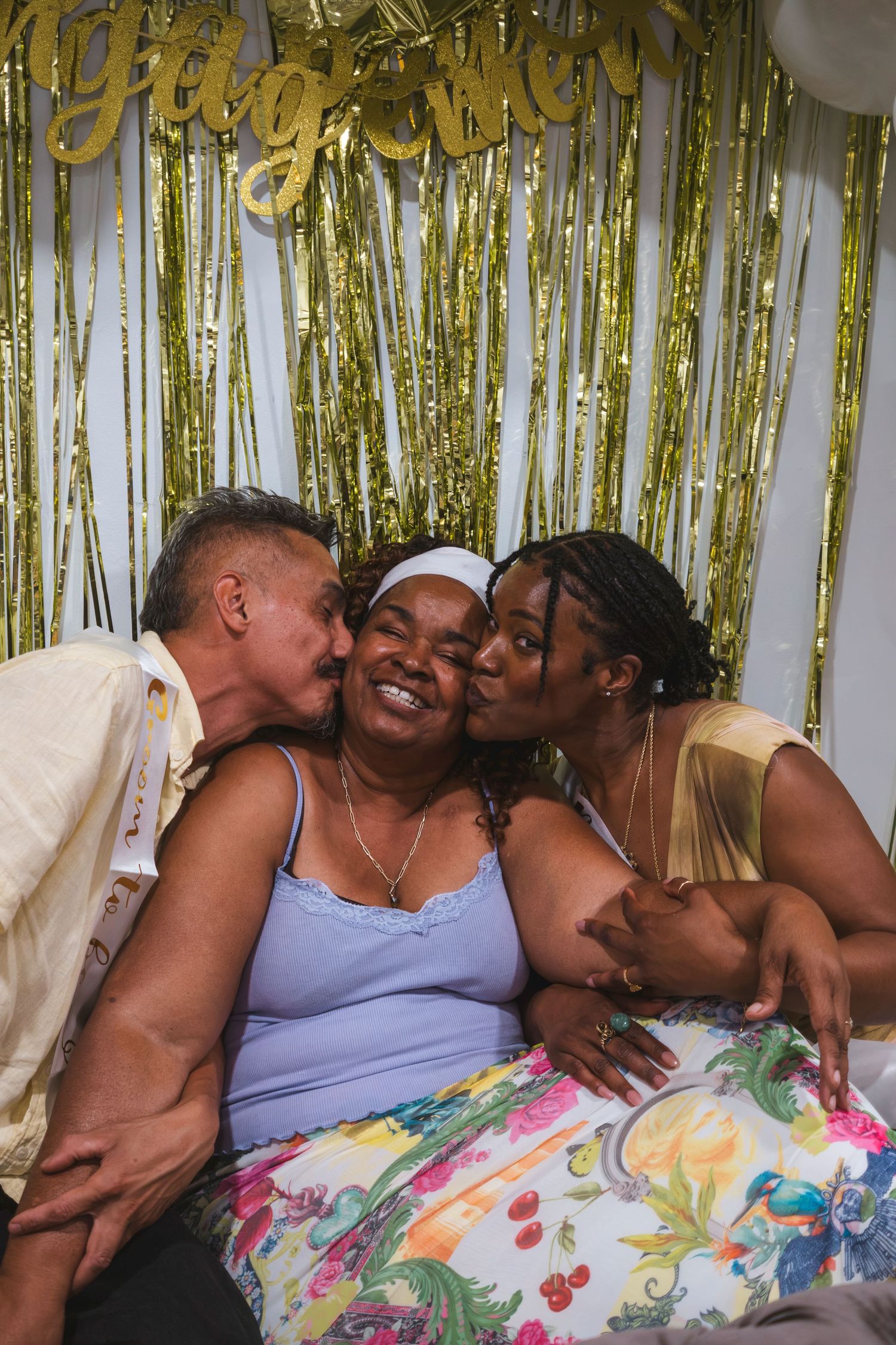 Two people kissing an older woman on the cheeks at a celebration with gold tinsel backdrop.