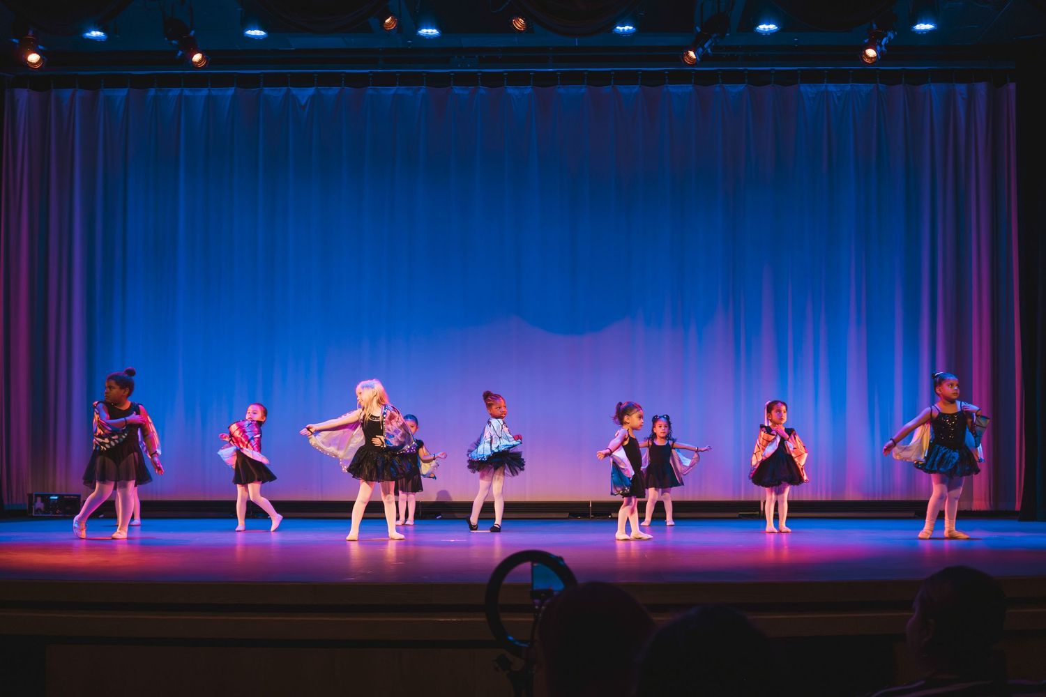 Young dancers perform on a lit stage with blue curtains during a children's dance recital.