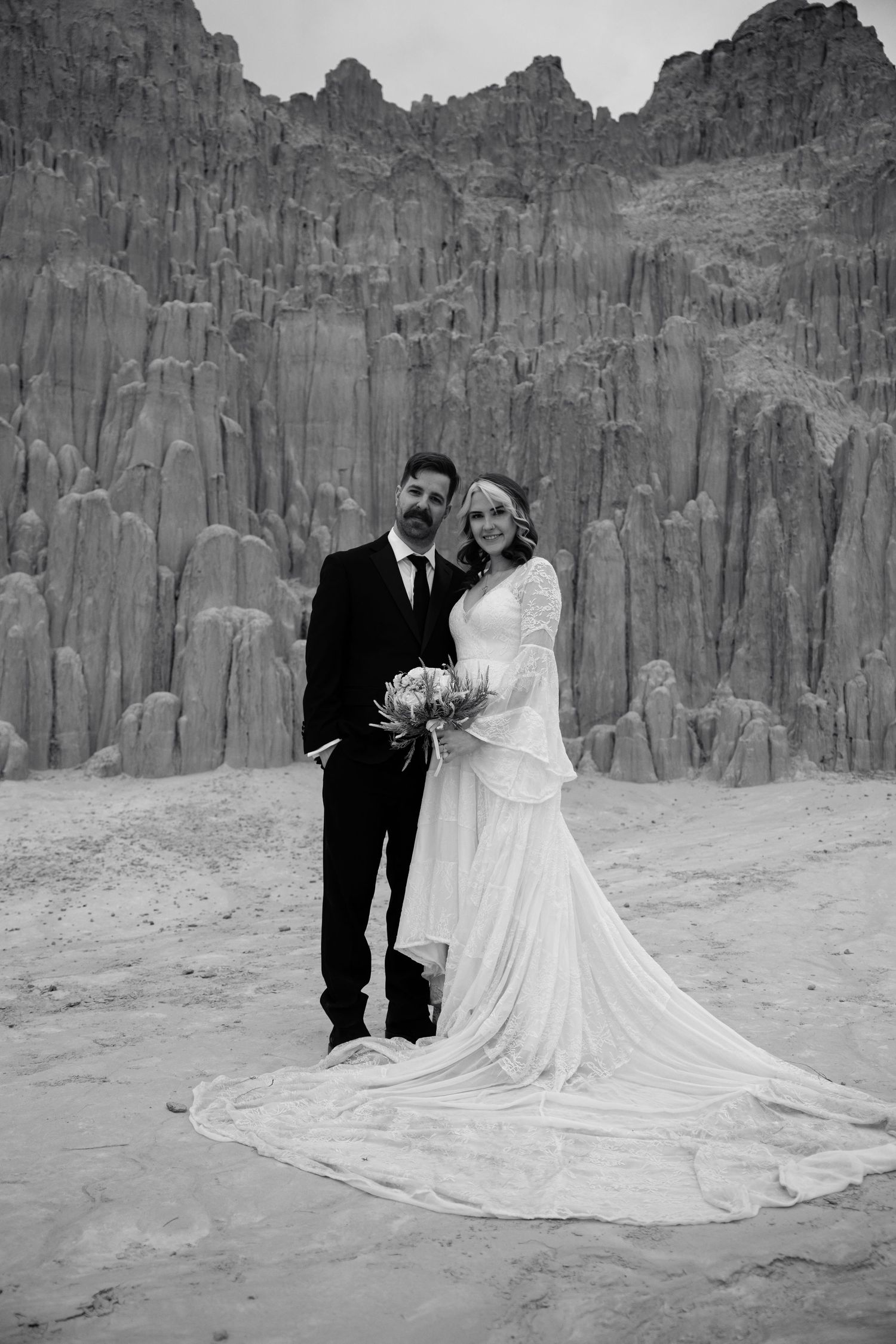 Black and white portrait of bride and groom standing together before dramatic desert rock spire formations.