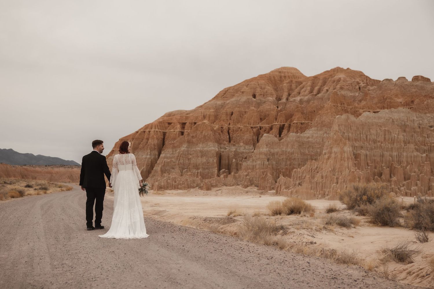 Bride and groom walk hand in hand along desert road toward dramatic sandstone rock formations.