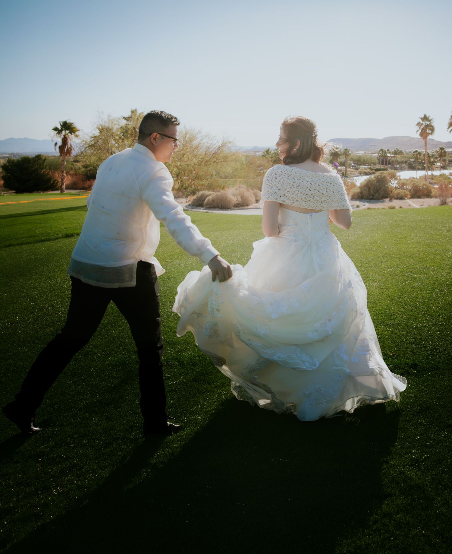Bride in white ball gown and groom in white shirt hold hands running across green lawn at sunset.
