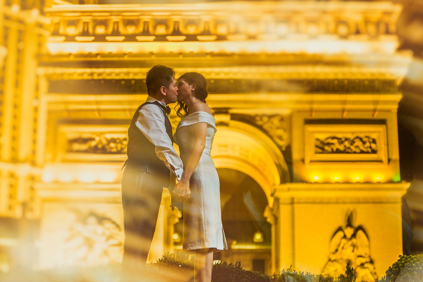 Couple sharing a romantic kiss in front of a glowing Arc de Triomphe replica at night.