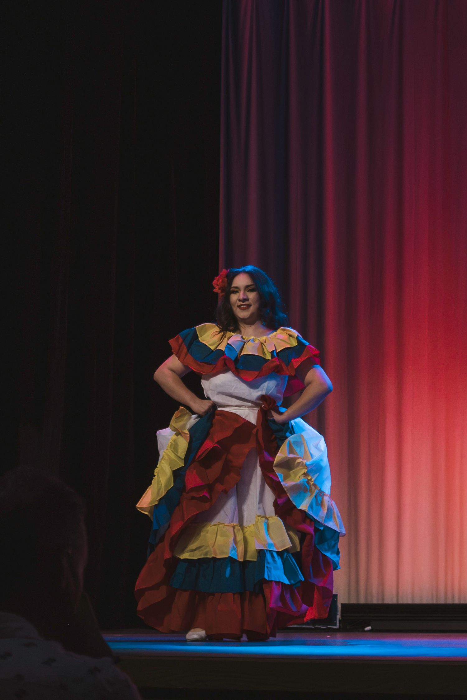 Woman performing traditional Latin dance in colorful ruffled dress on stage with red curtain backdrop.