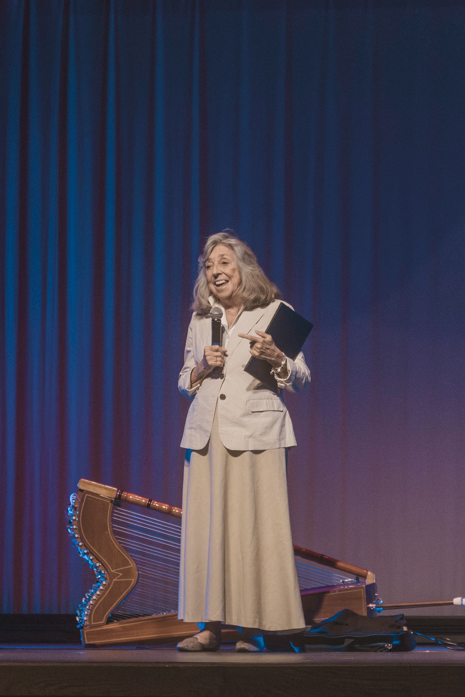Elderly woman in white suit speaking on stage with microphone, holding book near decorative harp.