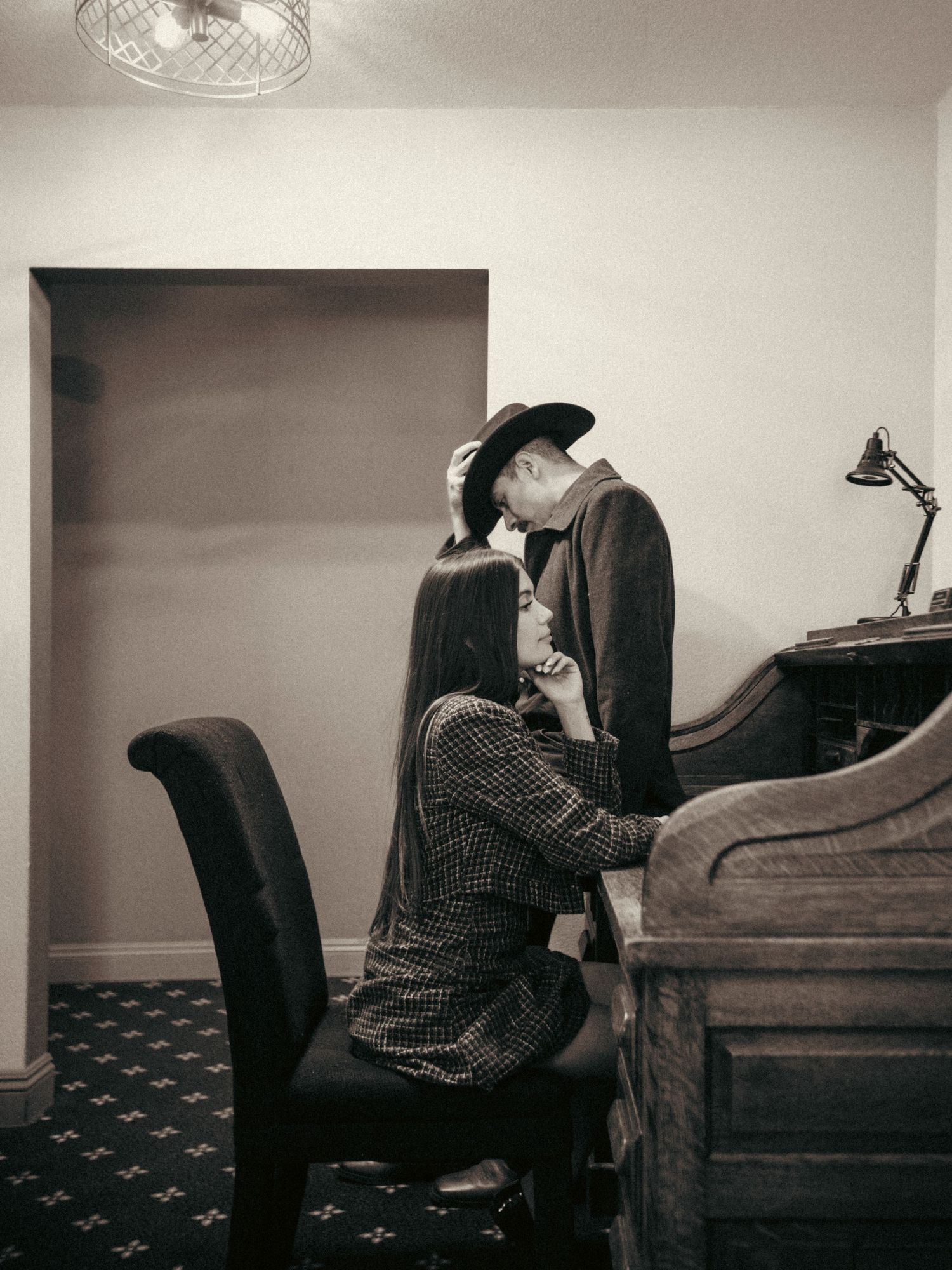 A couple shares a romantic moment near a vintage piano in a dimly lit room, captured in black and white photography.
