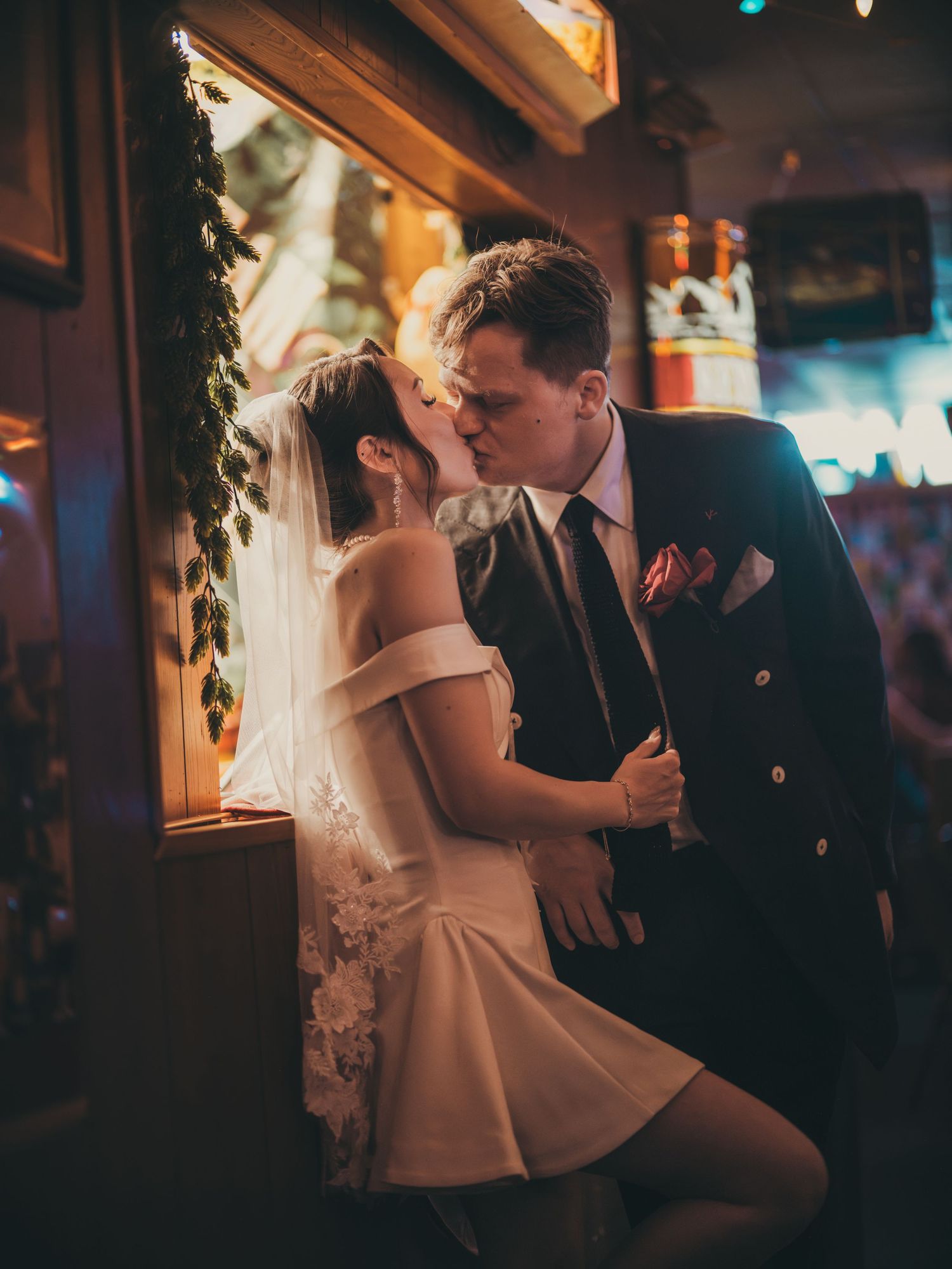 Bride and groom share a romantic kiss in a warmly lit venue, bride wearing a short white dress and veil.