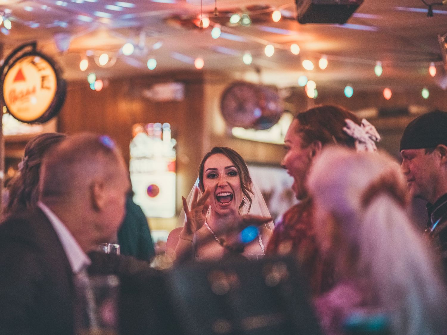 A woman laughs joyfully with friends at a warmly lit bar decorated with colorful string lights.