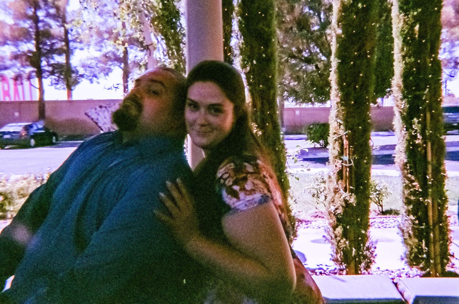 A couple poses together outdoors near decorative trees with warm string lights in a candid night photo.