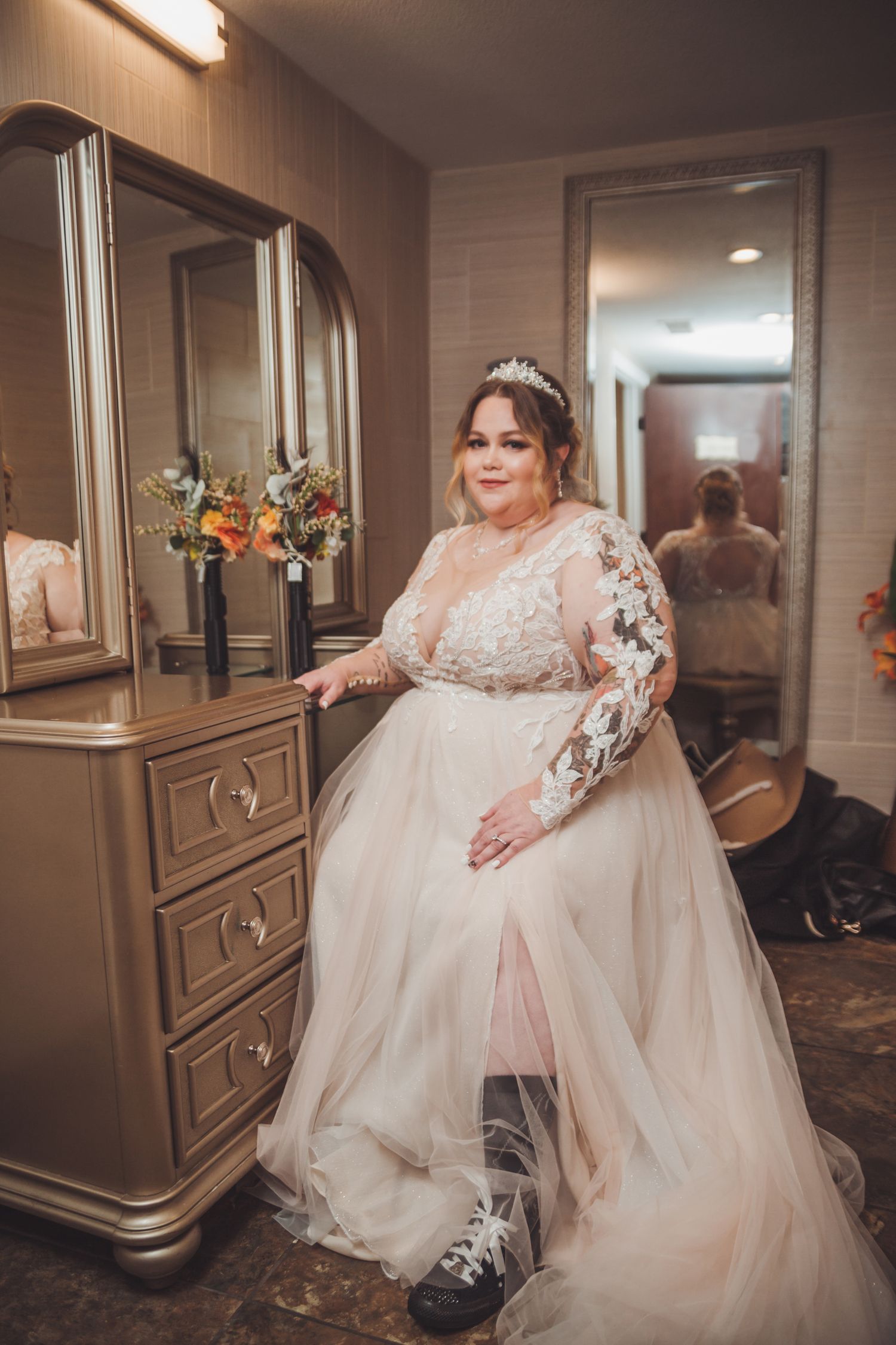Plus-size bride in lace long-sleeve wedding gown with tulle skirt poses by dresser with floral bouquet.