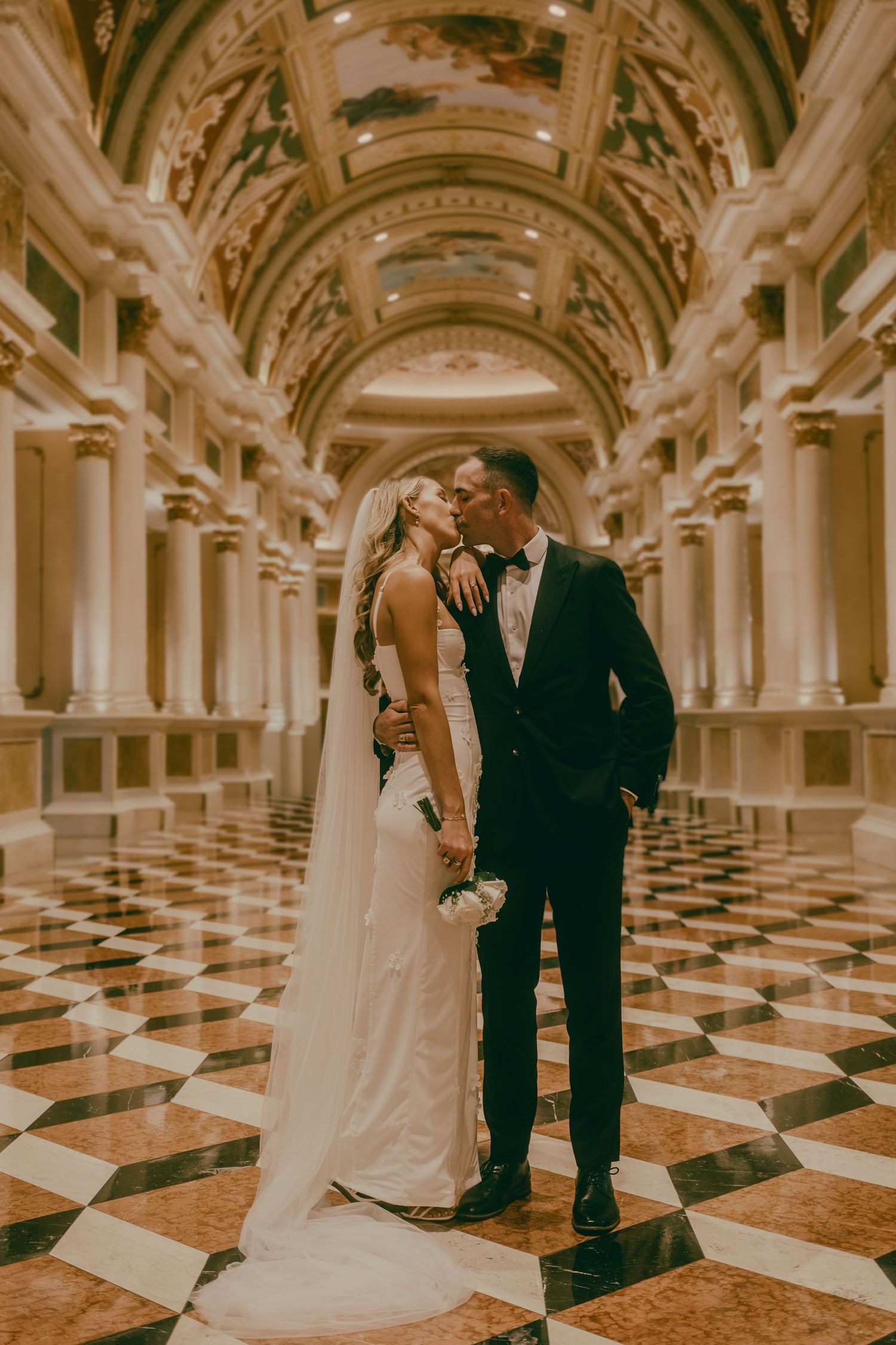 Wedding couple sharing a kiss in a grand ballroom with checkered marble floors.