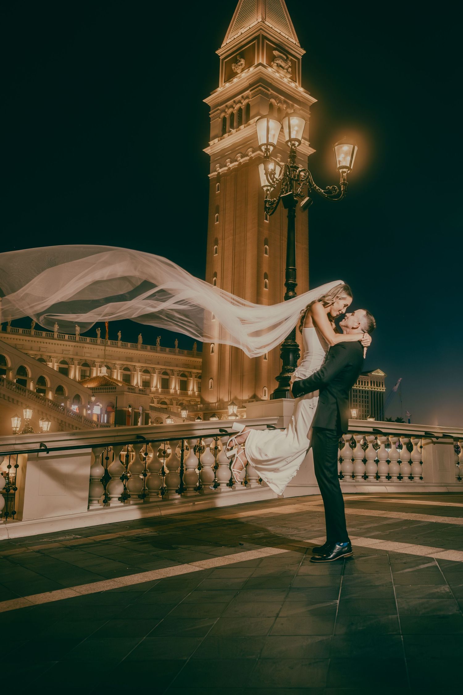 Groom lifting bride with flowing veil near The Venetian tower at night in Las Vegas.