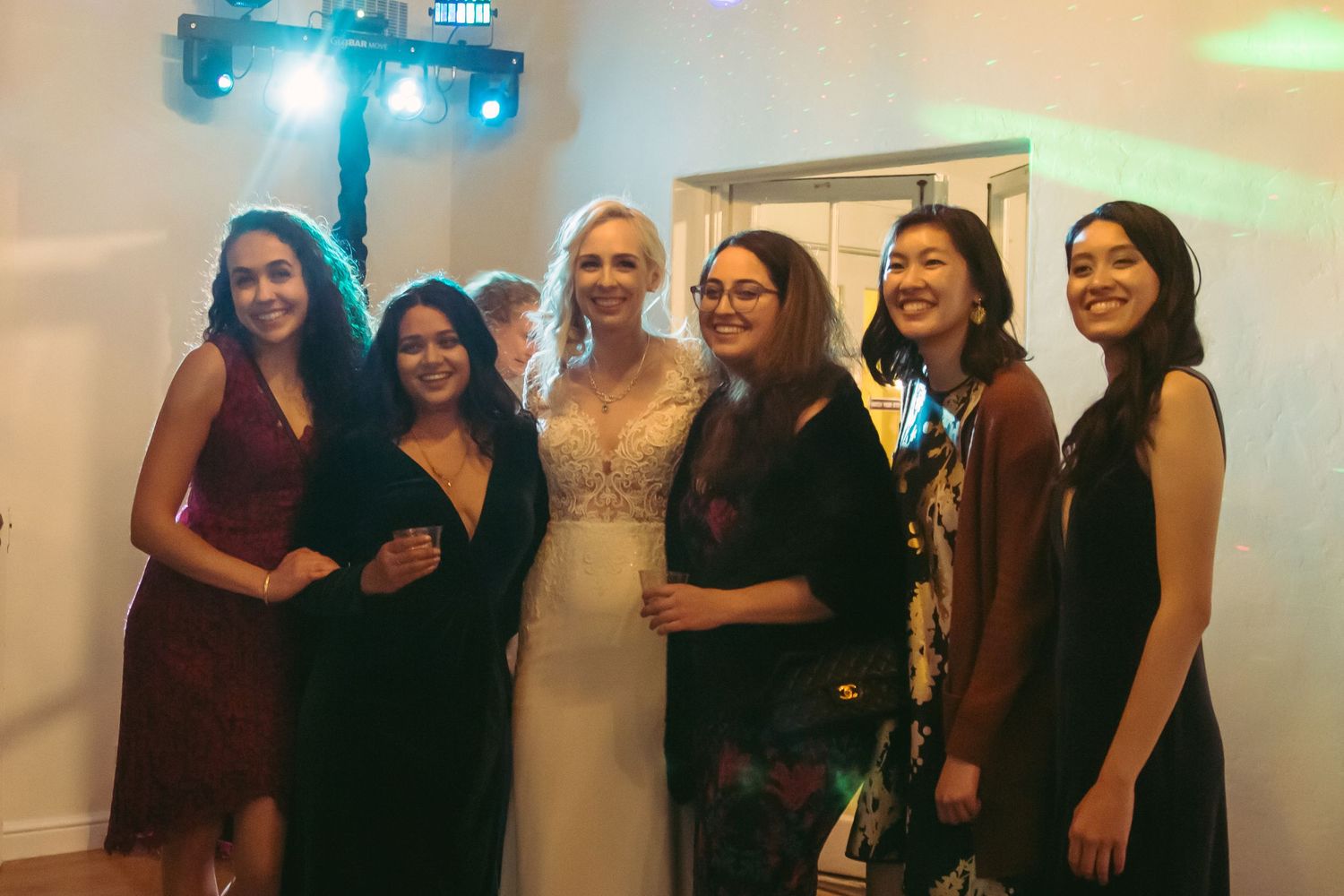 Bride in lace gown posing with six female guests under disco lights at wedding reception.