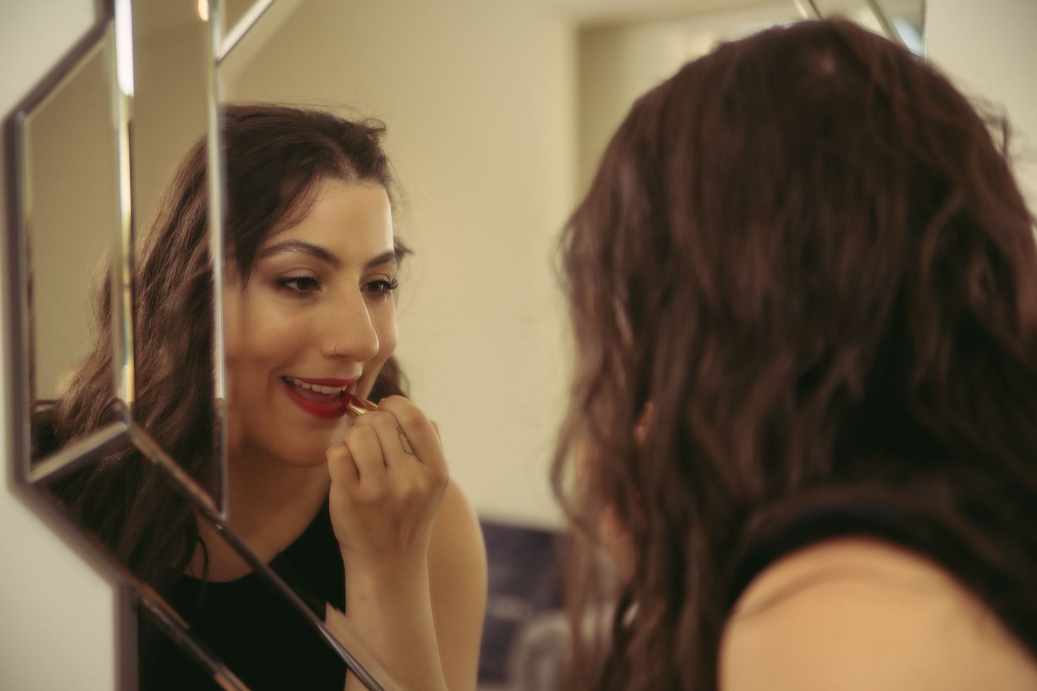 A smiling woman applies red lipstick while looking into a small square mirror.