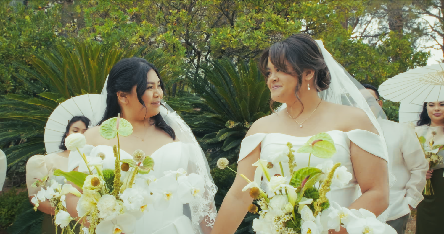 Two smiling brides in white gowns and veils share a joyful moment holding white bouquets.