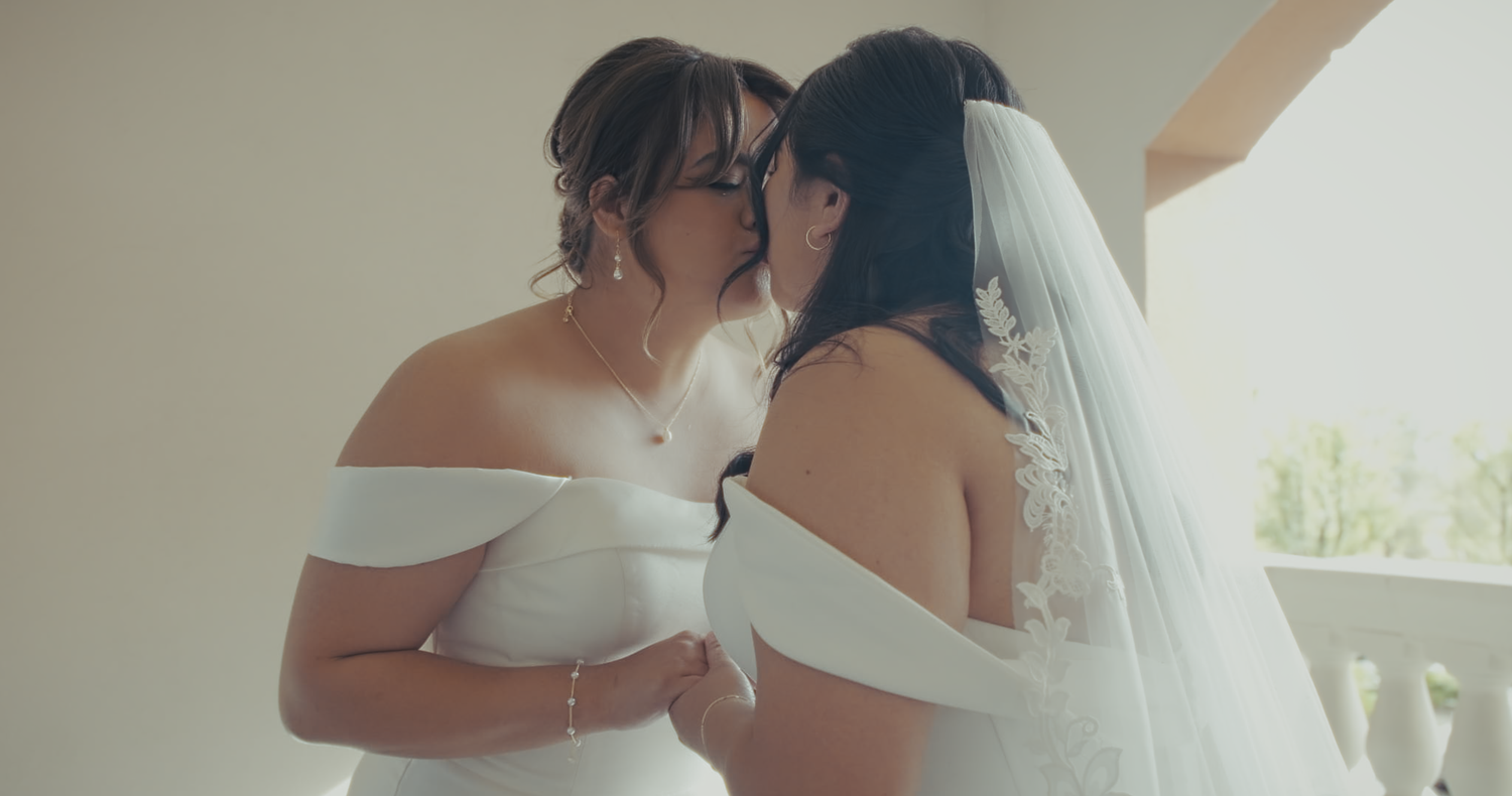 Two brides in white off-shoulder gowns share a tender kiss indoors.
