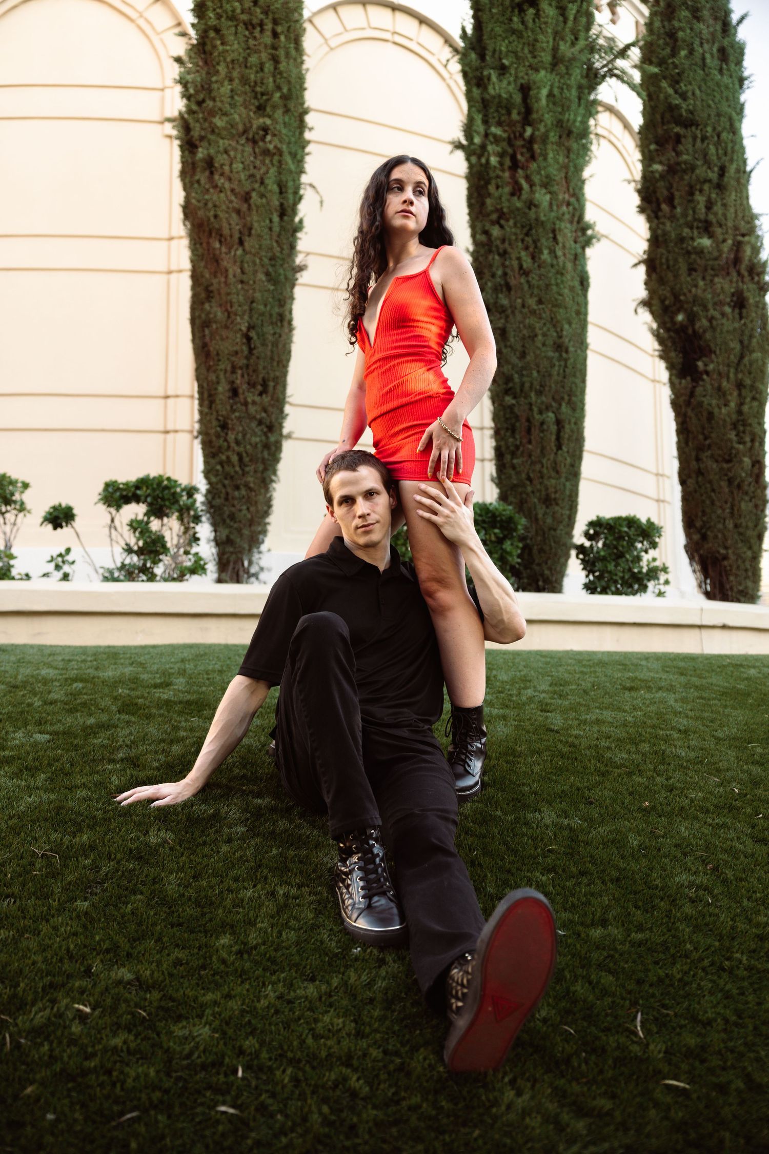 Couple posing on green lawn, woman in red mini dress standing behind seated man in black outfit.