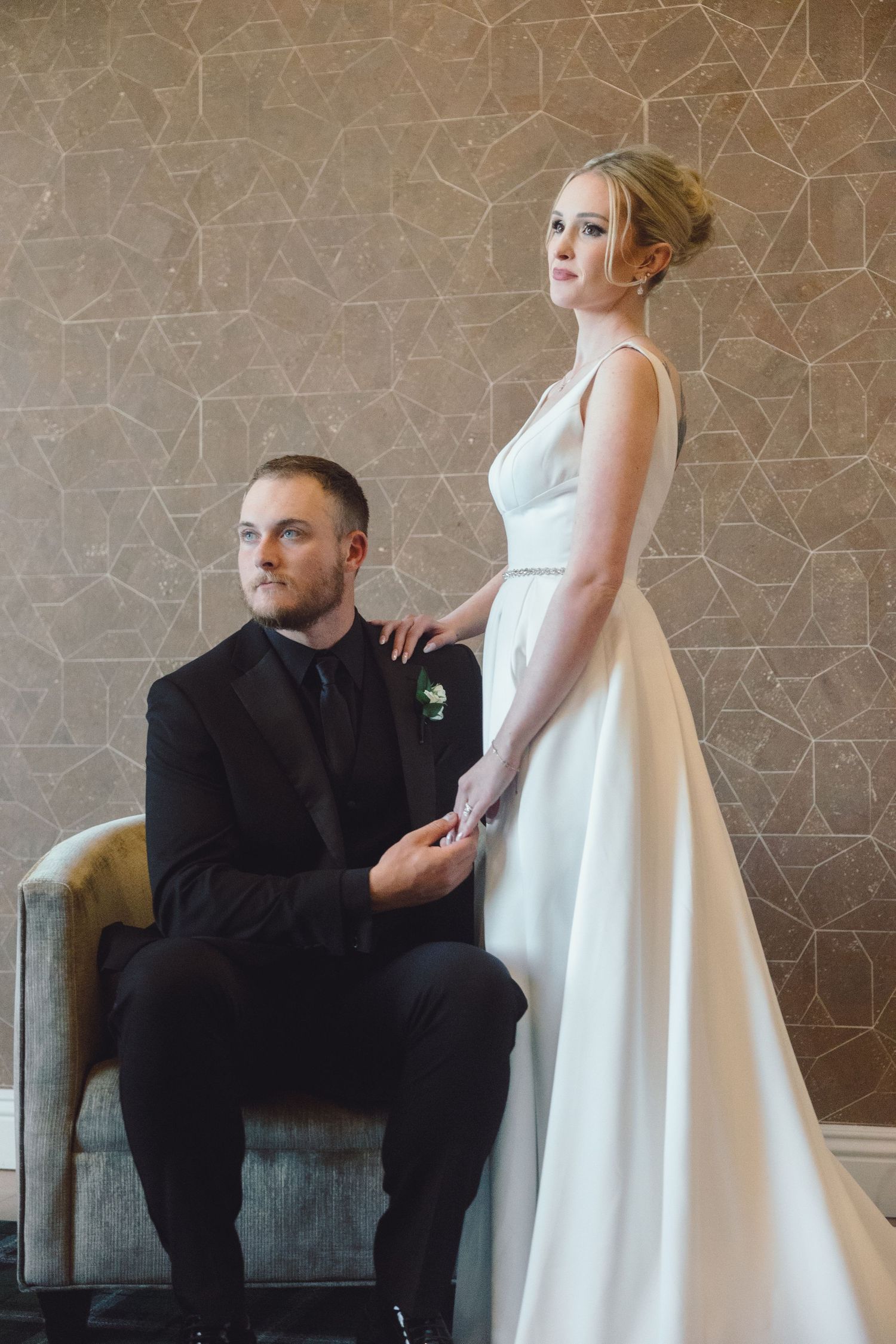 A bride in a white gown stands beside a seated groom in a black suit in an elegant interior setting.