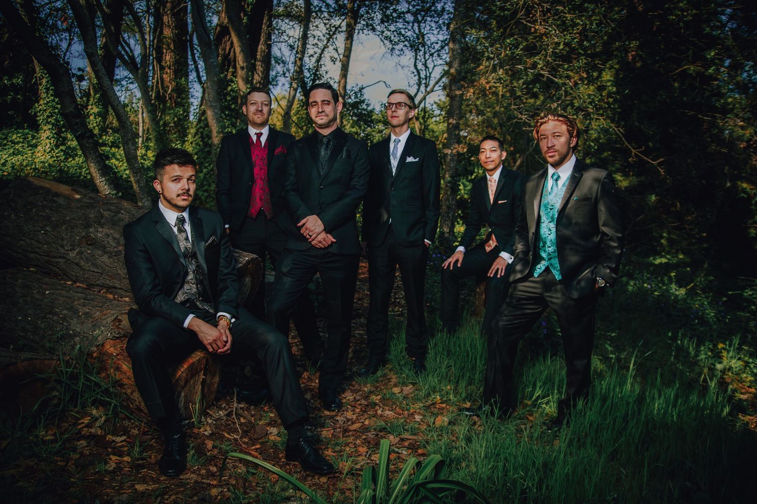 Six men in formal suits pose outdoors in a lush green forest setting for a wedding party photo.