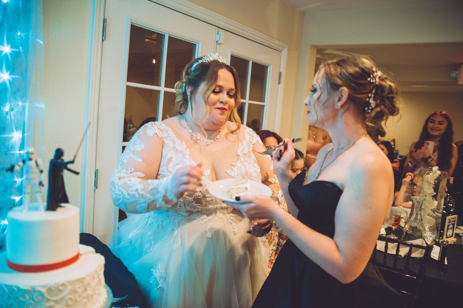 Bride in lace gown laughing as bridesmaid feeds her wedding cake at indoor reception with Star Wars topper.