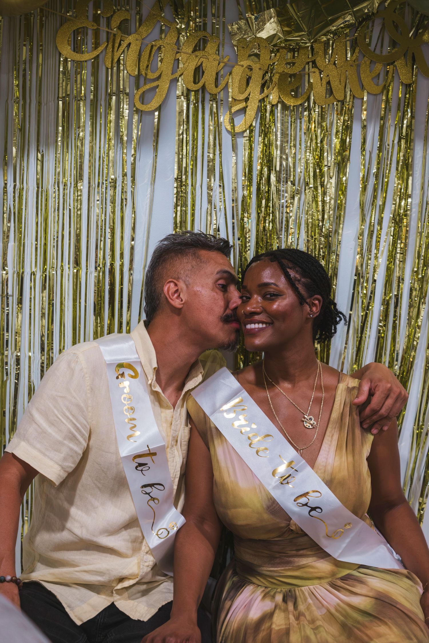 Couple wearing engagement sashes kissing at a gold glitter engagement party celebration backdrop.