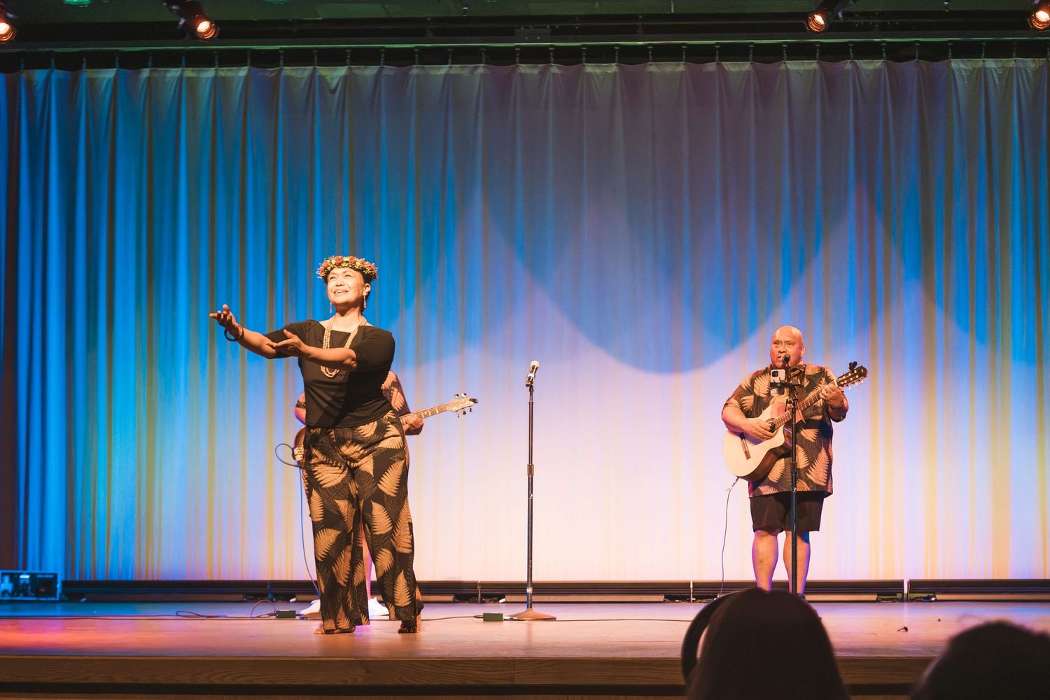 Two musicians perform on stage, one singing and one playing guitar, in front of a blue curtain backdrop.
