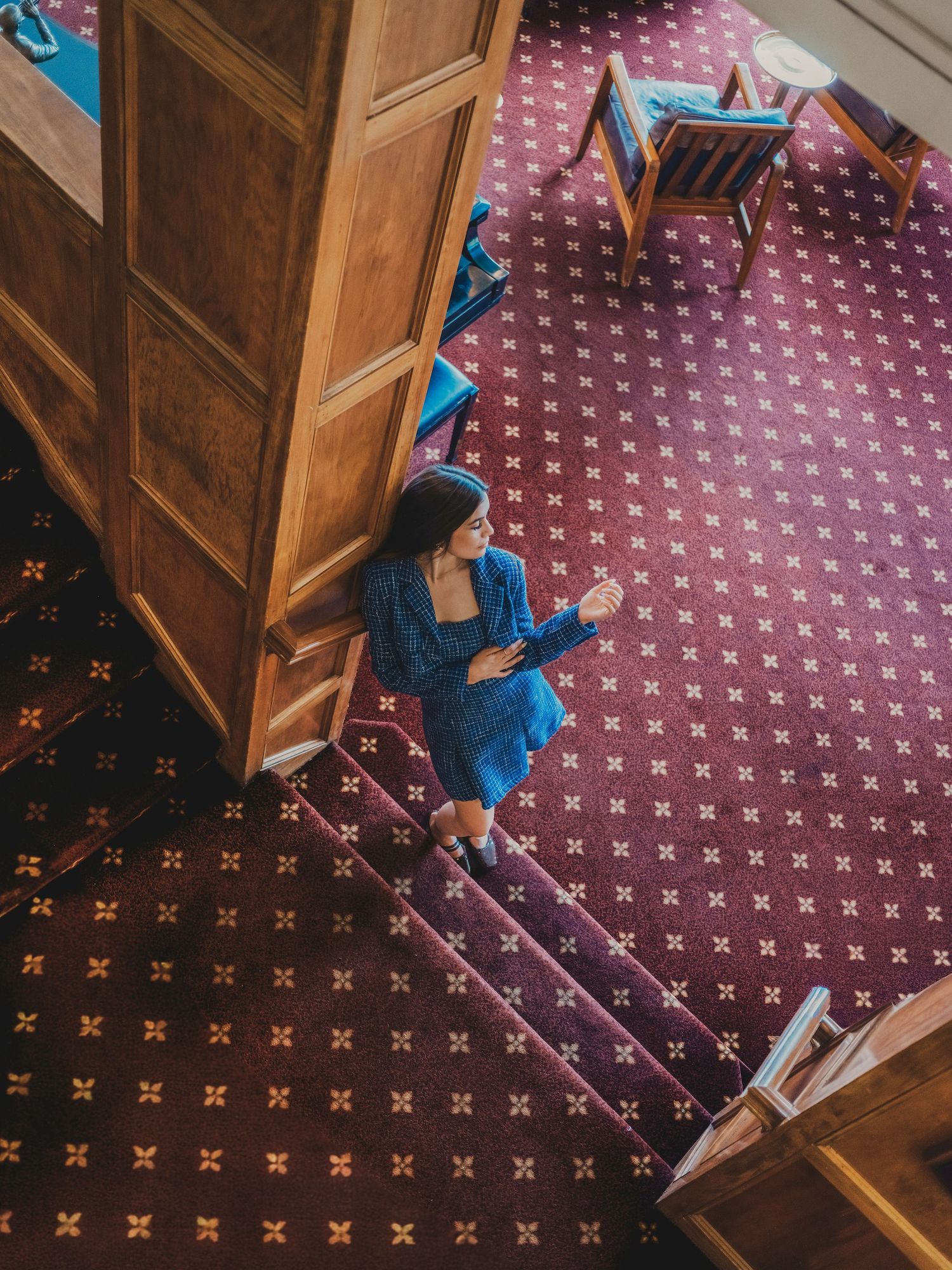 Aerial view of a woman in a blue dress on patterned red carpet stairs in an elegant hotel interior.