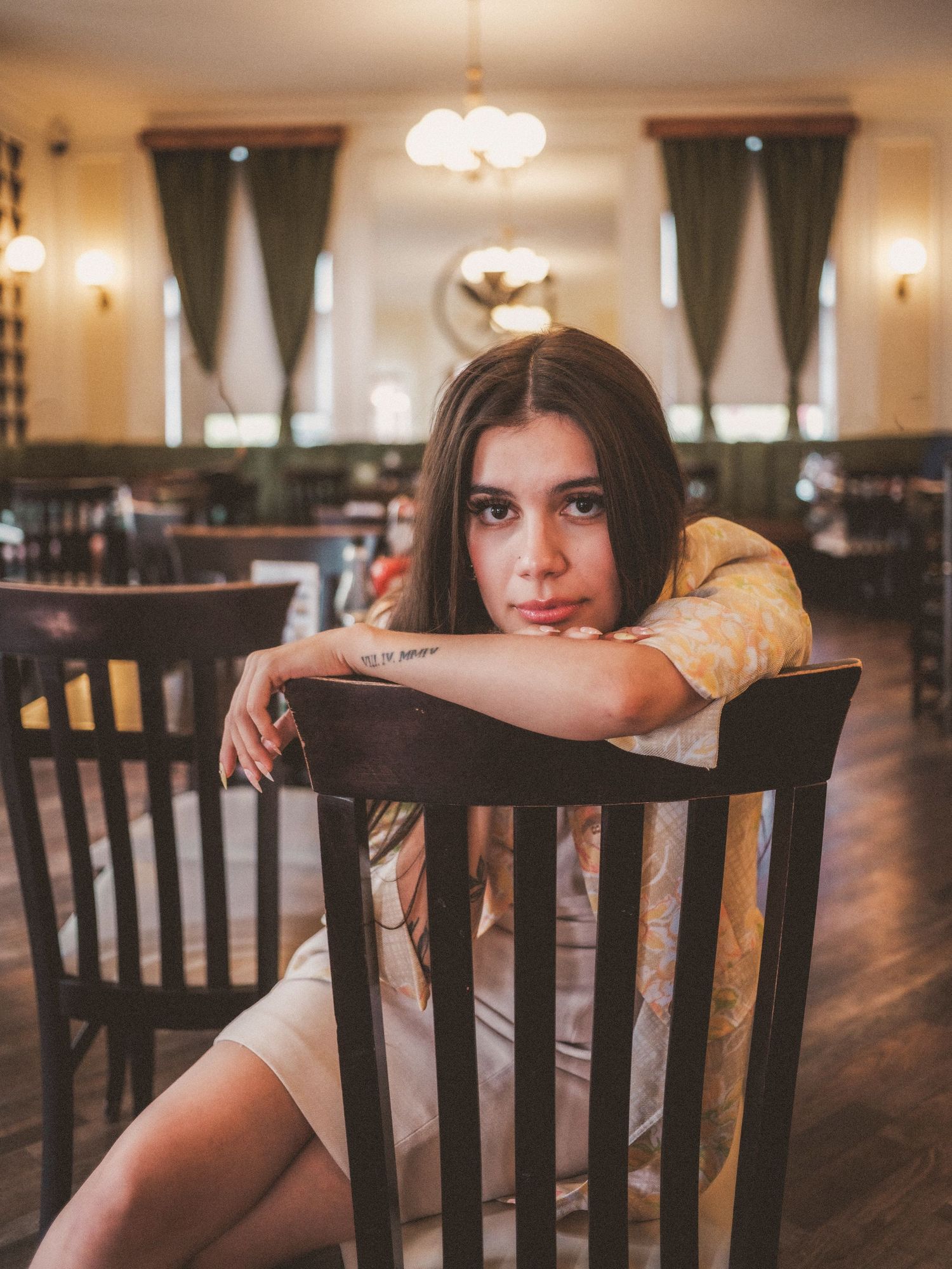 Young woman with tattoo sits backwards on wooden chair in elegant restaurant interior.