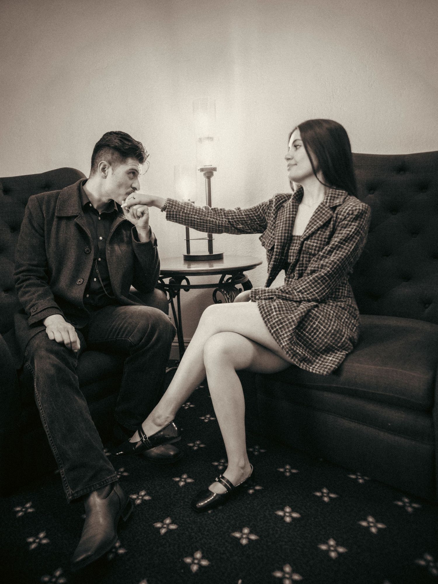 Black and white photo of a couple sitting on a sofa, woman extending hand to man in vintage-style setting.