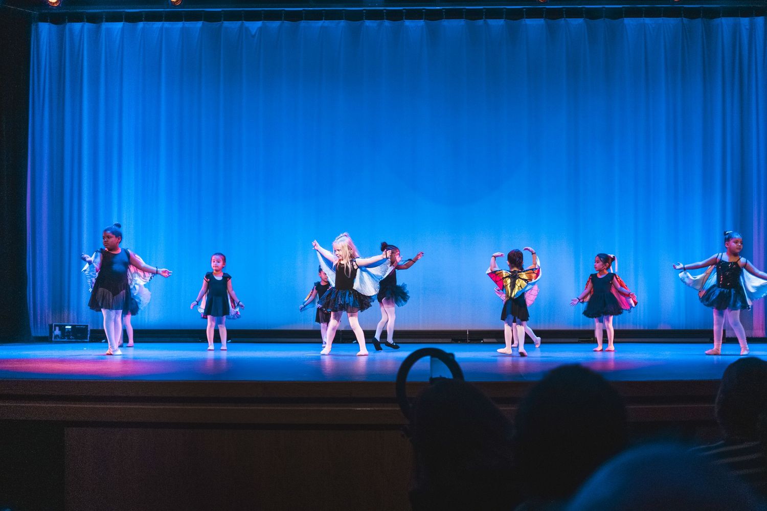 Young girls in black tutus perform a dance recital on a blue-lit stage during a children's ballet show.