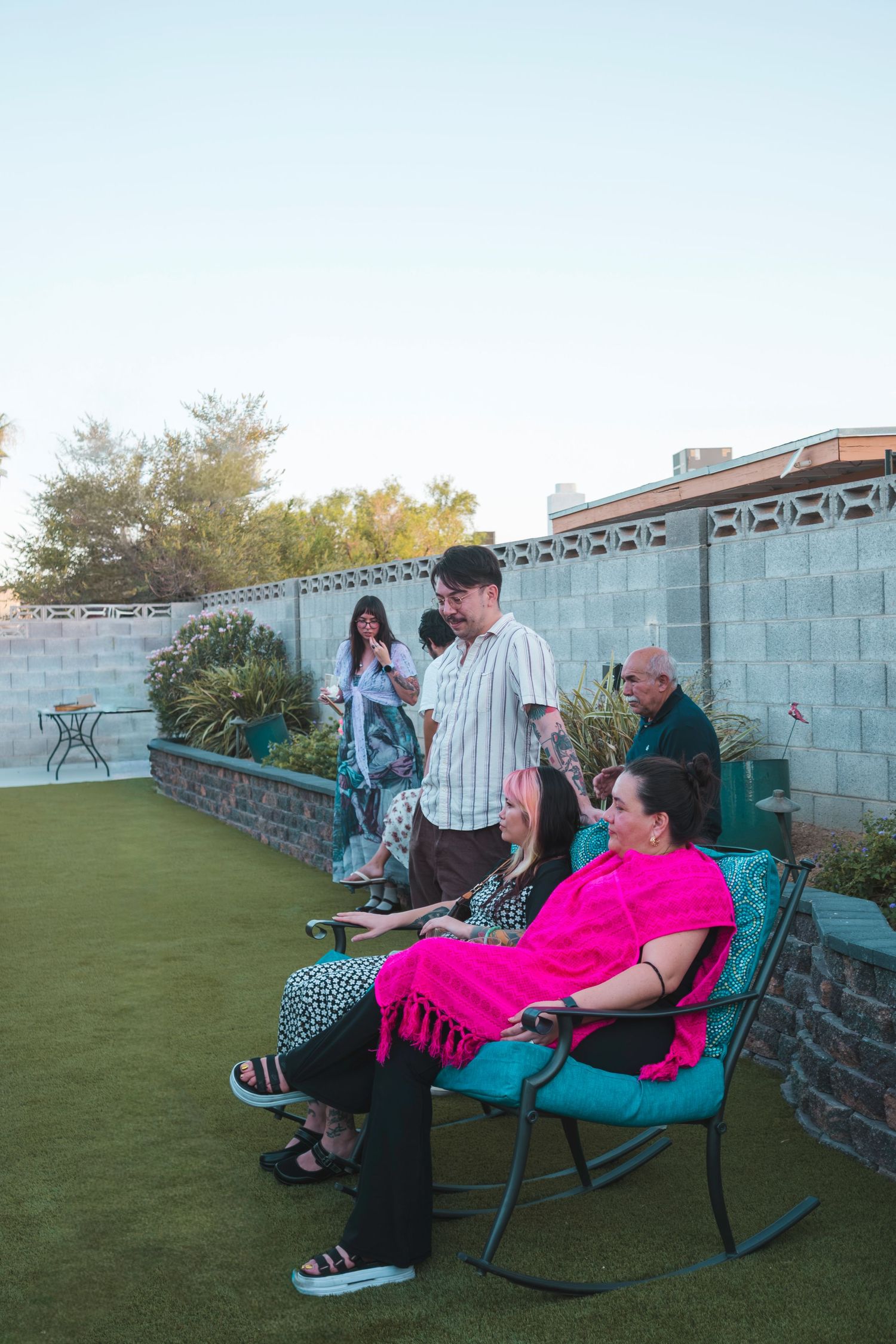 Group of people enjoying an outdoor backyard gathering at dusk, with a woman in pink relaxing in a rocking chair.