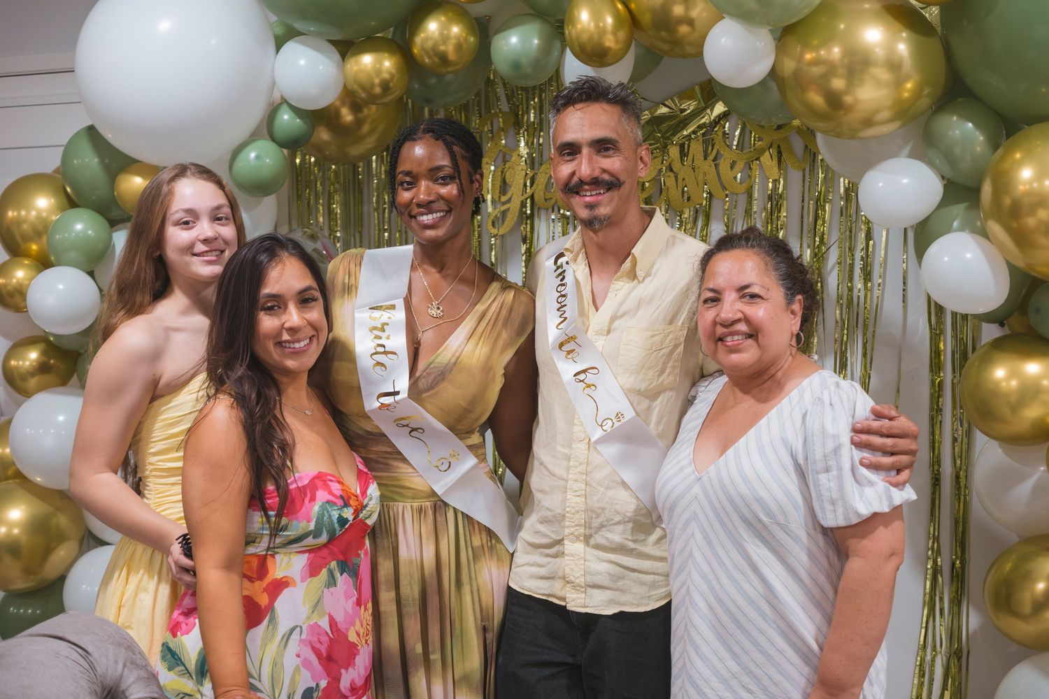 Group of five people posing together at a festive event with green and gold balloon arch backdrop.