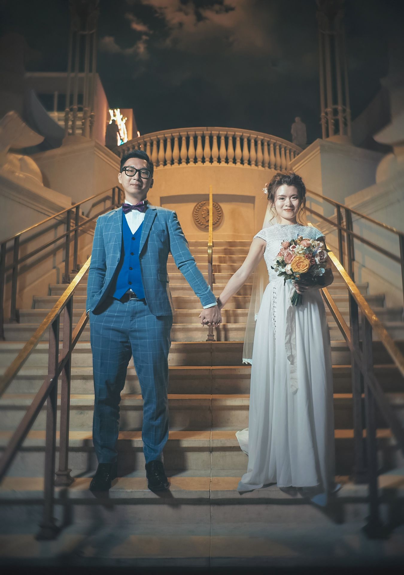 Couple in wedding attire holding hands on grand staircase, groom in blue suit, bride in white gown holding bouquet.