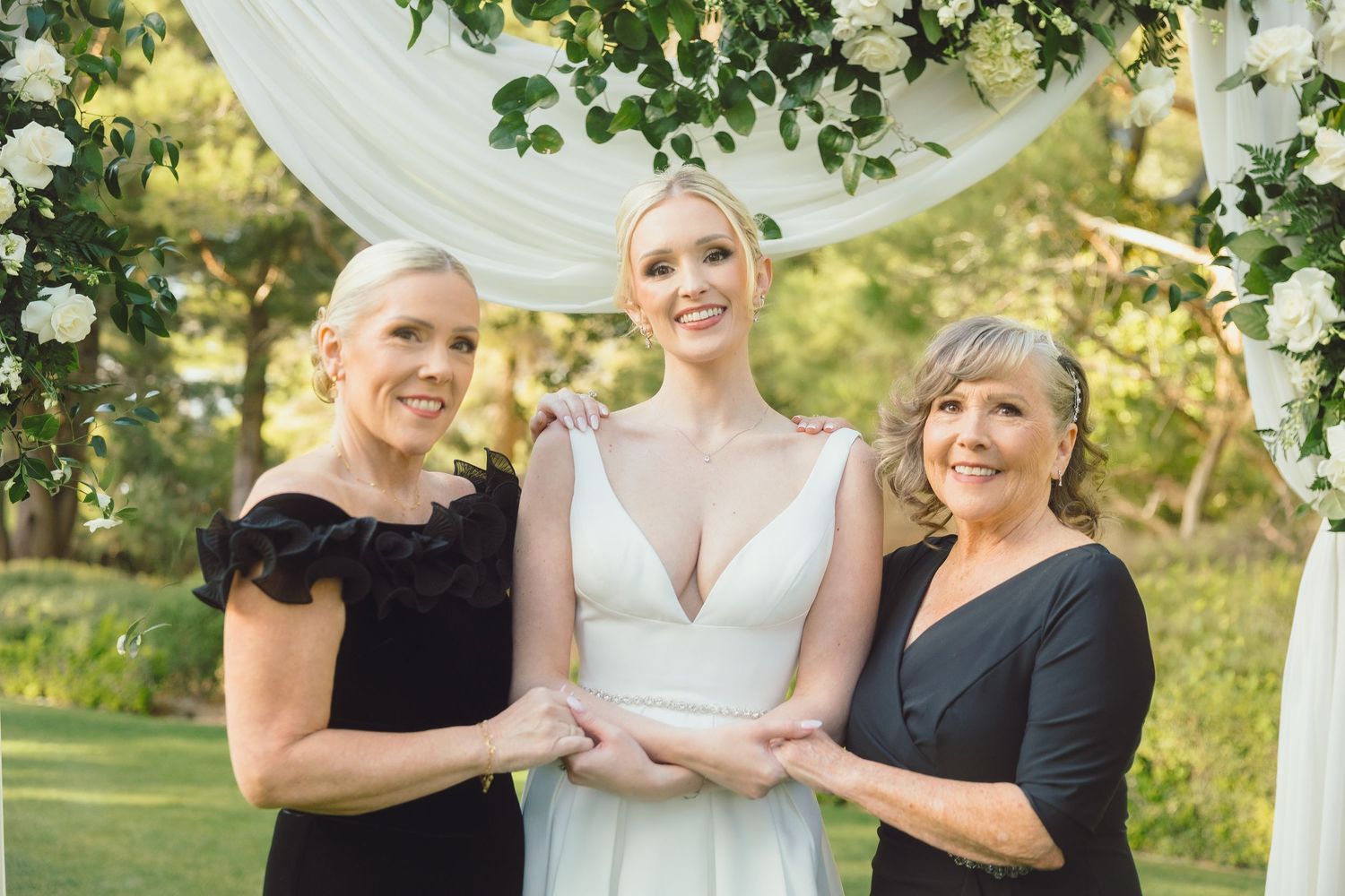 Bride posing with two guests in black dresses under greenery wedding arch.