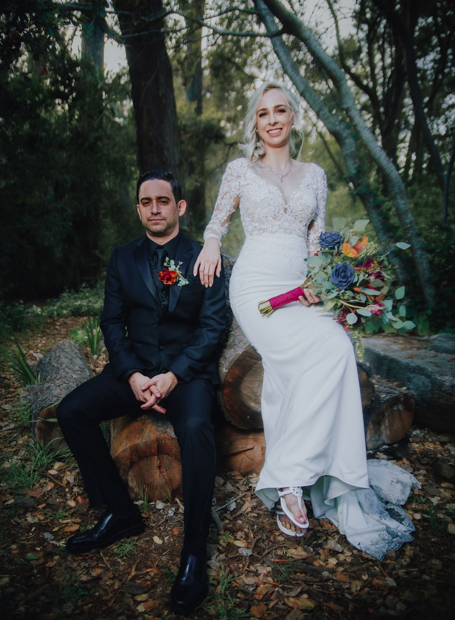 Bride in white lace gown holding colorful bouquet sits beside groom in black suit on log in forest setting.