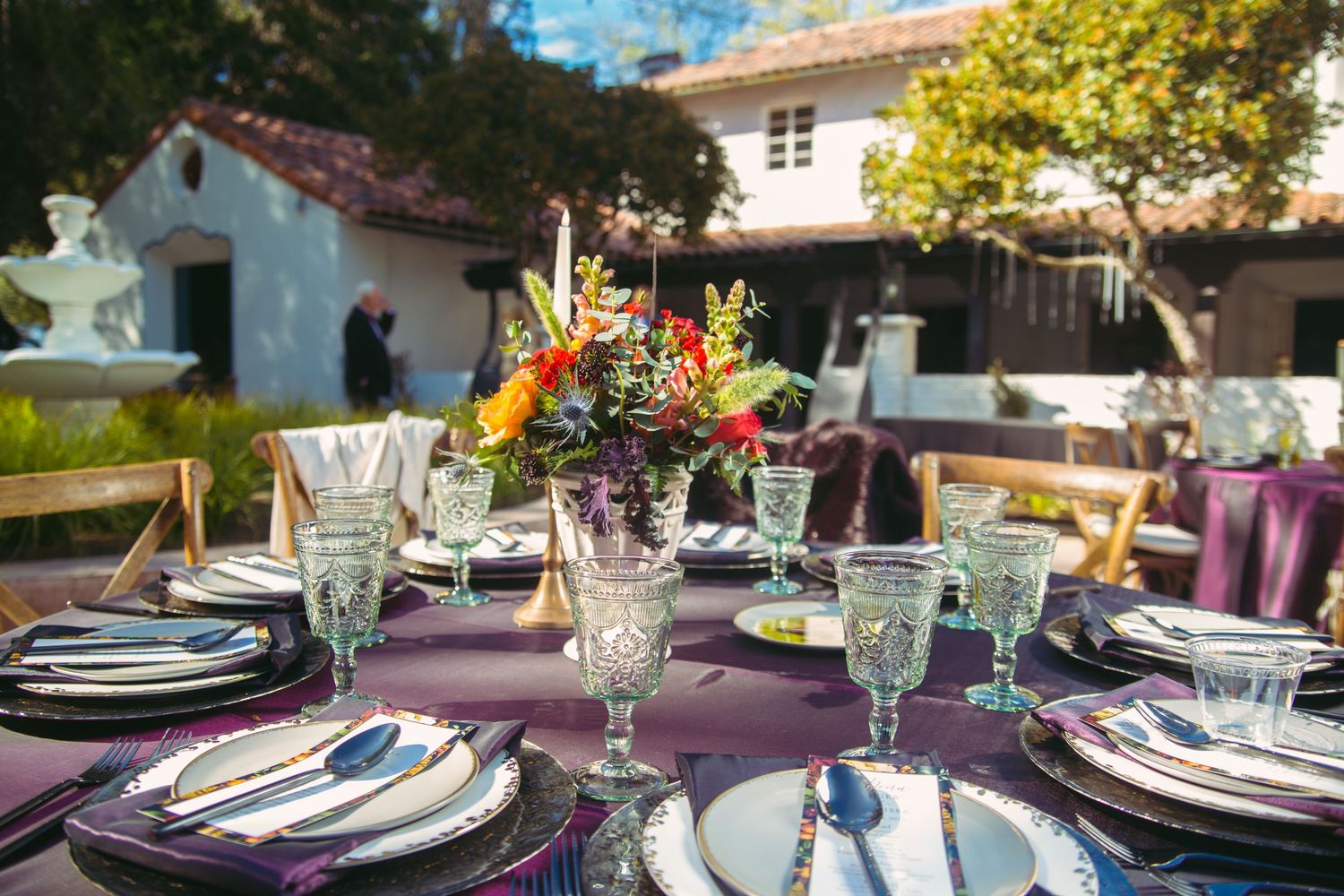 Elegant outdoor wedding reception table set with purple linens, crystal glasses, and colorful floral centerpiece.
