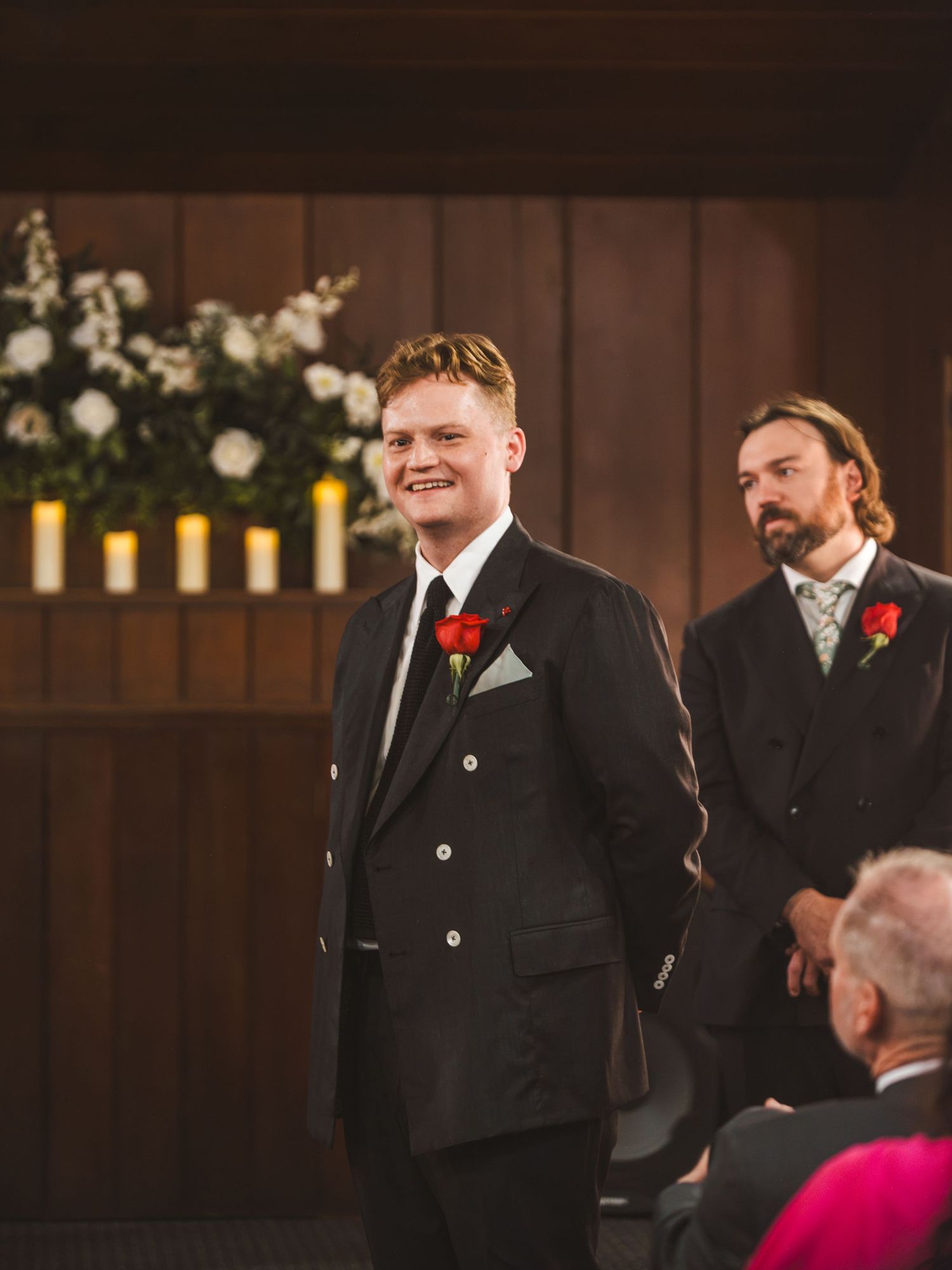 Groom in black suit with red rose boutonniere smiling at wedding altar with groomsman standing behind him.