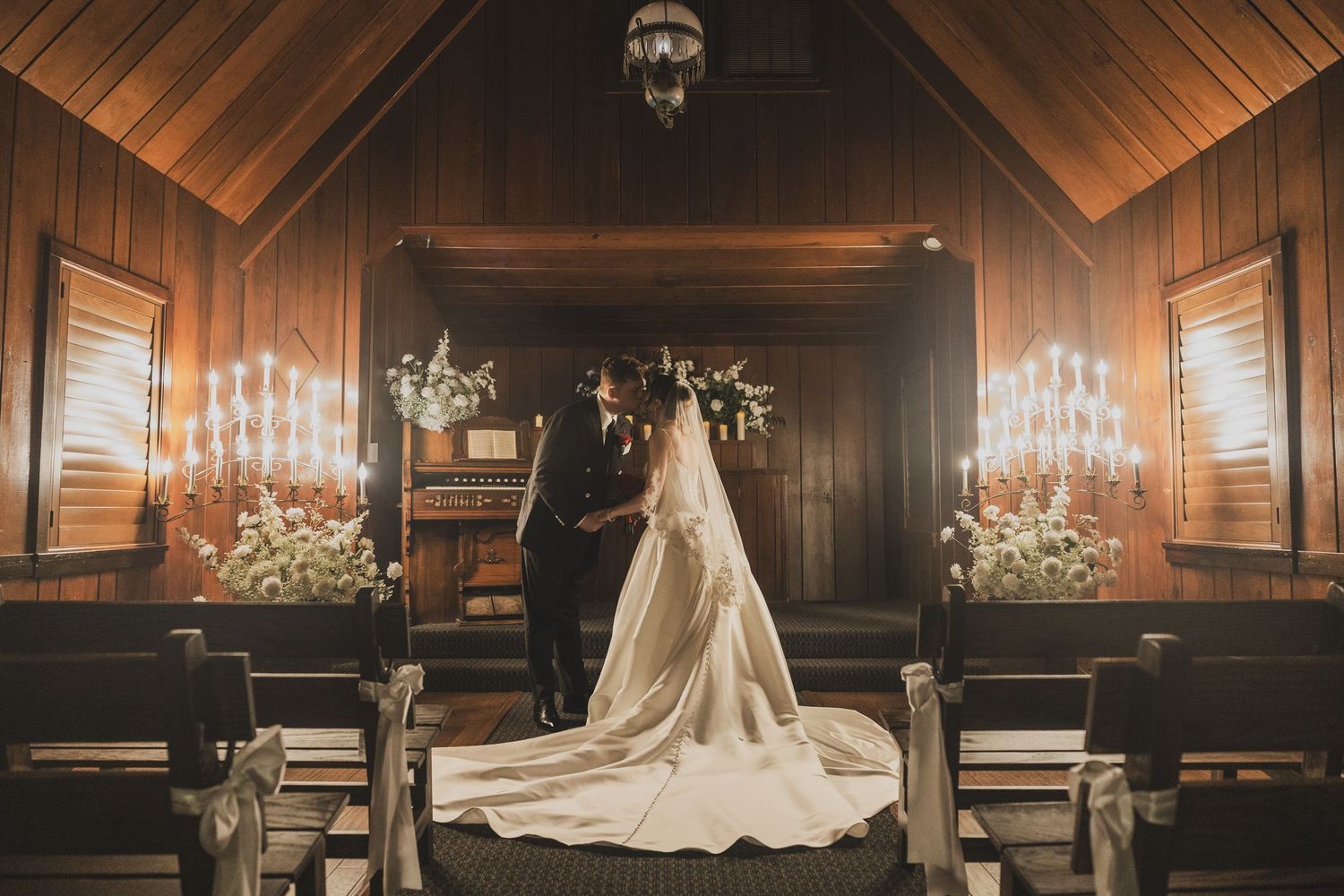 Bride and groom exchange vows at a candlelit wooden chapel altar with floral arrangements.
